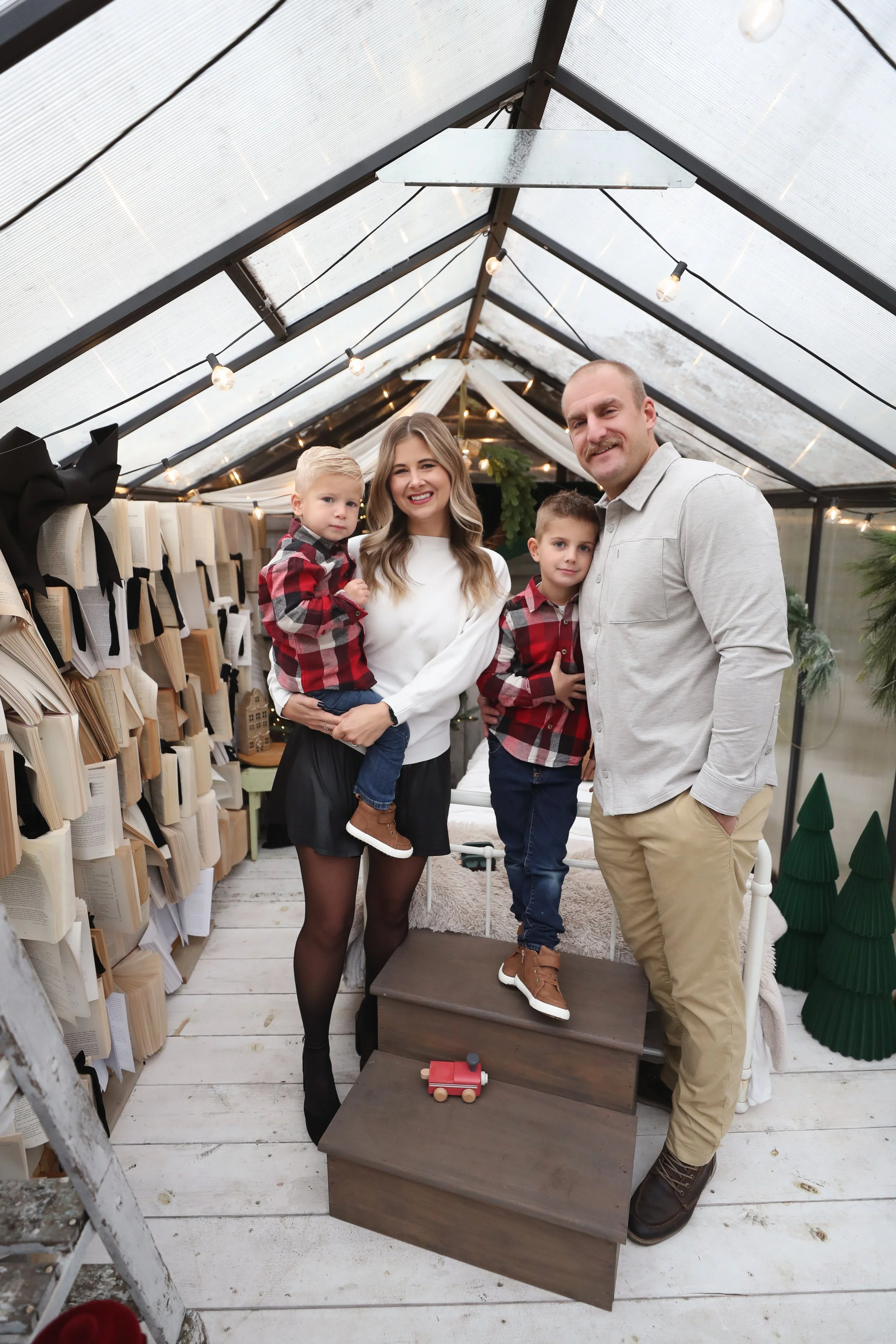 A family of four, parents and two children, standing inside a decorated greenhouse with books, Christmas trees, and ornaments, smiling at the camera during holiday time.