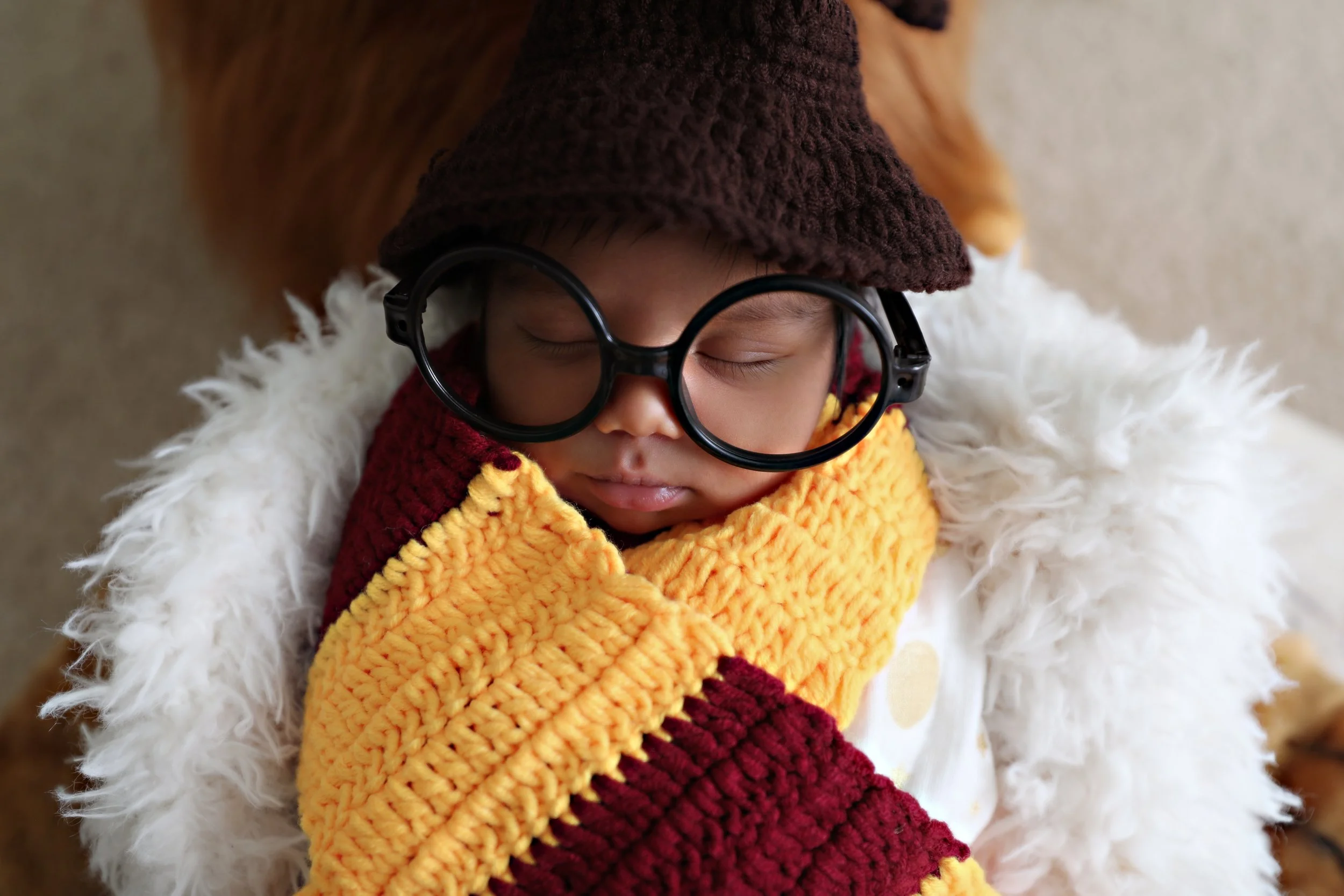 A young child dressed in winter clothing with a furry coat, colorful knitted scarf, glasses, and a knit hat, sleeping peacefully with eyes closed.