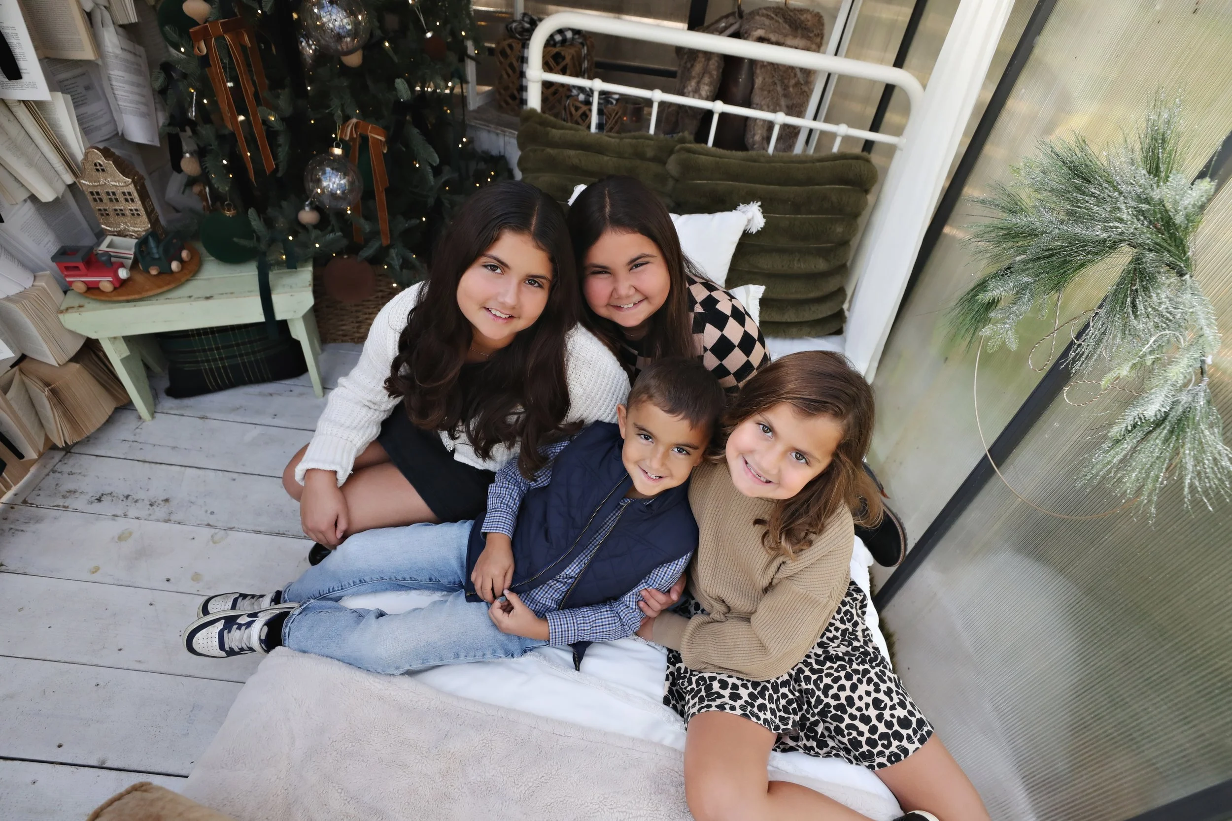 Four children, two girls and two boys, sit together on a bed in a cozy, decorated room with Christmas decorations, including a Christmas tree with ornaments and ribbon, and a small side table with decorative items.