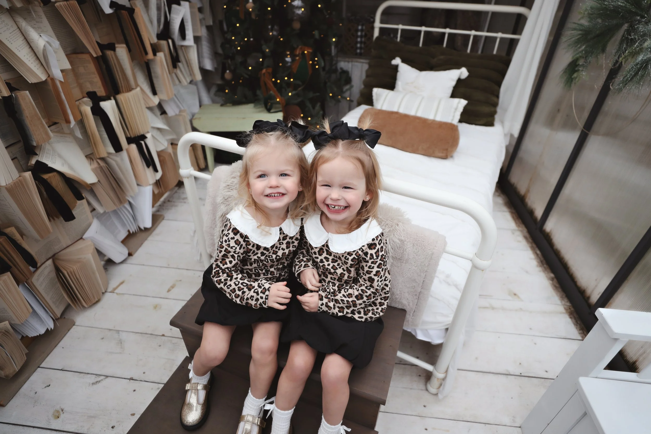Two young girls sitting together on a small table in a cozy room decorated for Christmas, with a decorated tree, bookshelves, and a bed with pillows and a blanket in the background.
