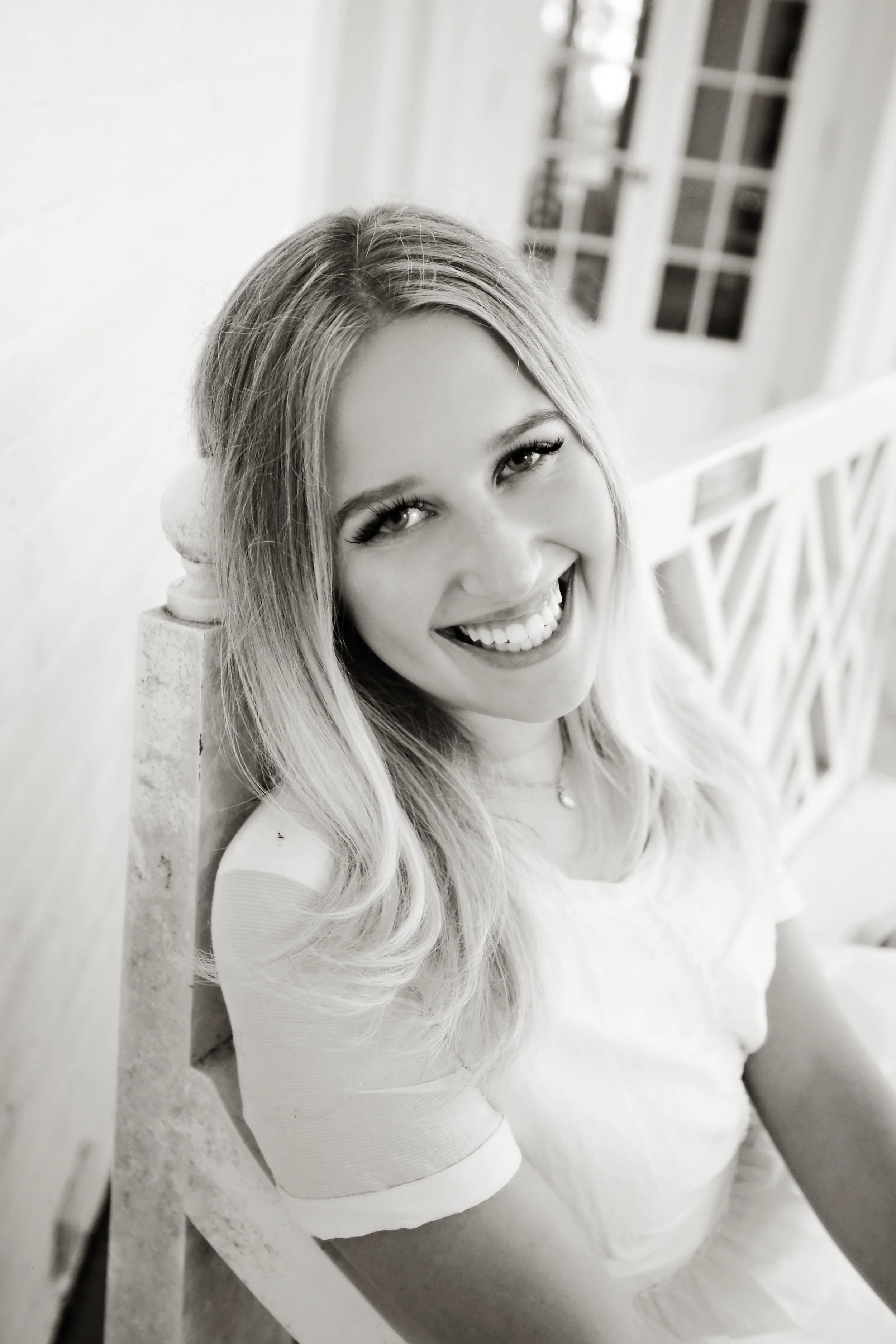 Black and white photo of a young woman with light wavy hair sitting on a bench, smiling brightly at the camera, indoors near windows.