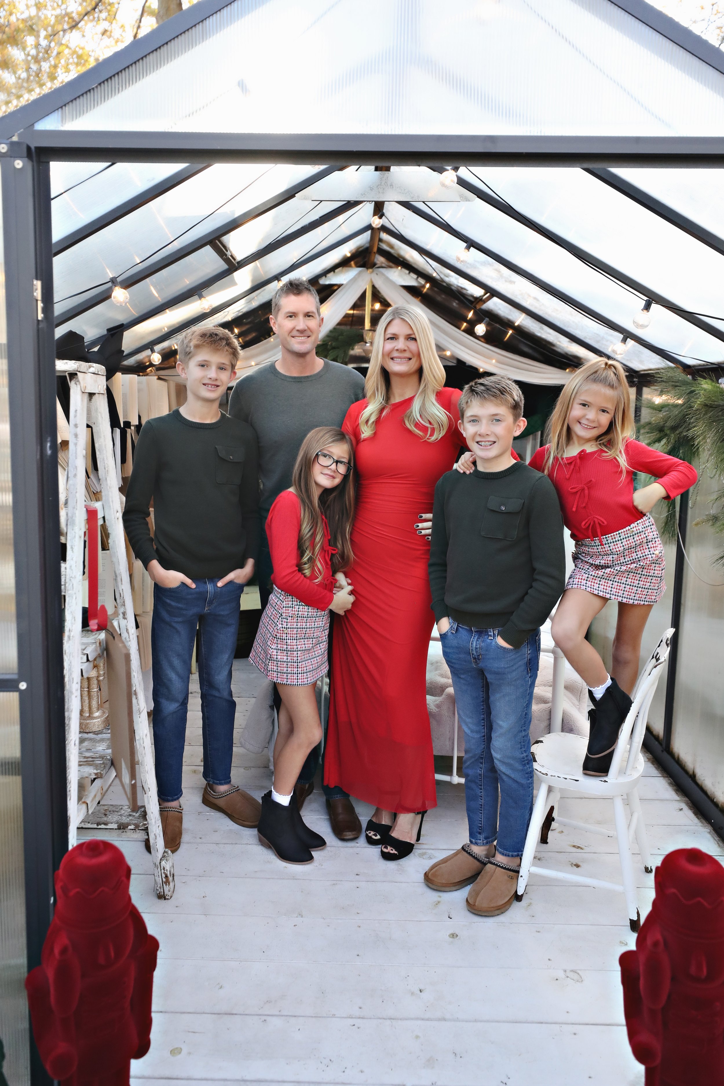 Family of six posing inside a greenhouse with string lights, decorated for the holiday season, wearing red and black outfits, with Christmas trees seen in the background.