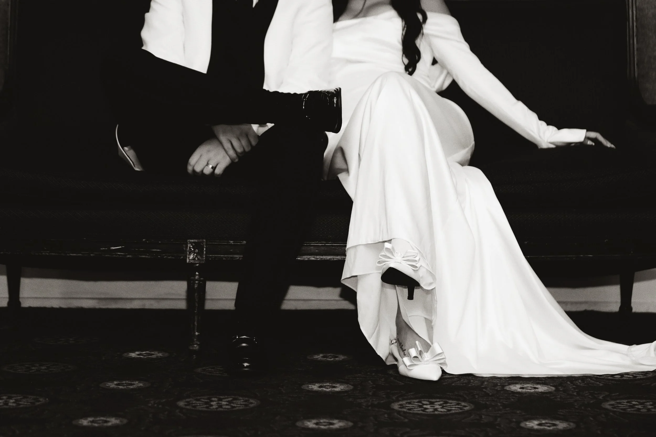 A black-and-white photo of a newlywed couple sitting on a vintage couch, with the bride wearing a long white wedding gown and showcasing a shoe with a bow, and the groom dressed in a dark suit, holding hands.
