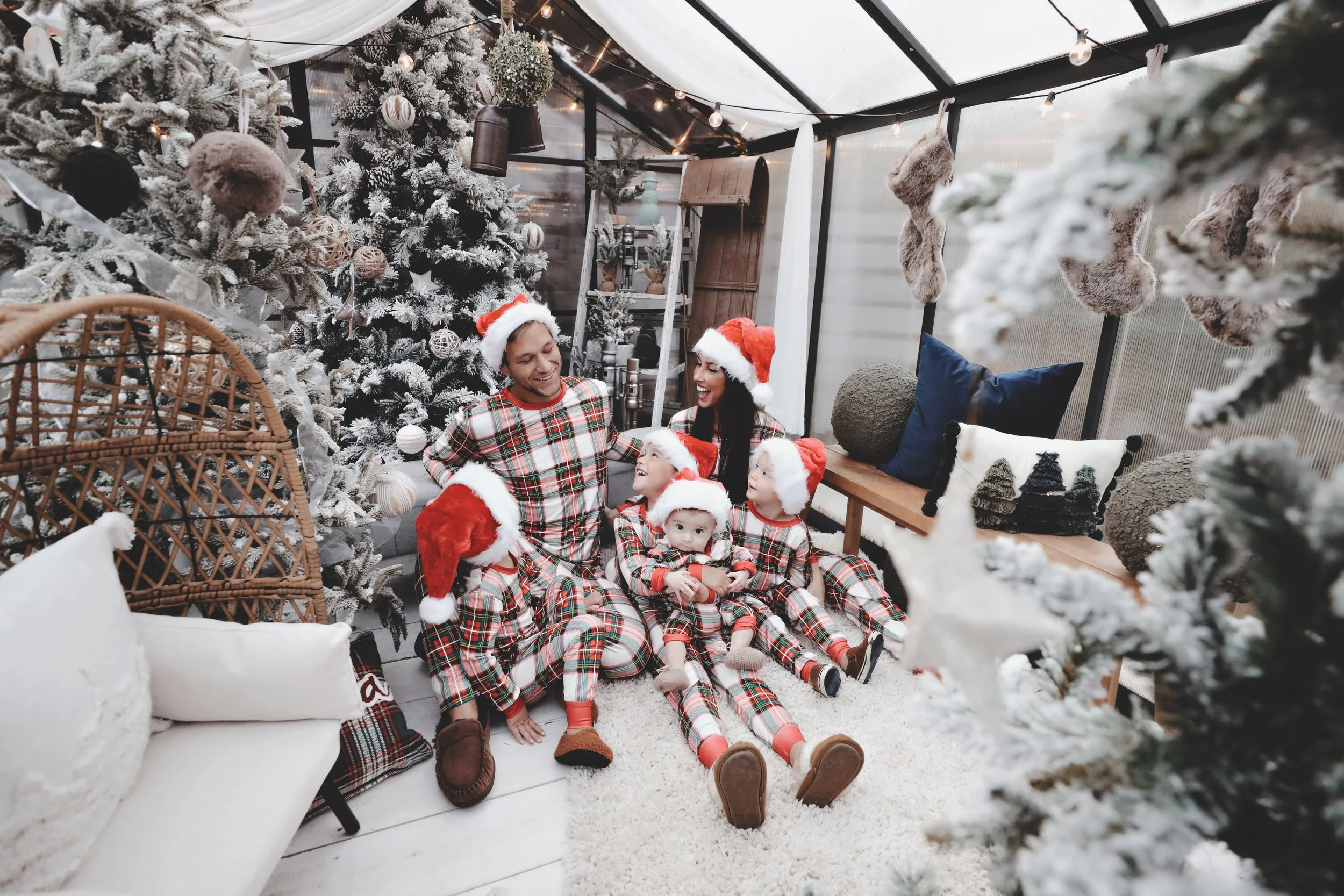 A family of five dressed in matching plaid pajamas and Santa hats sitting together in a decorated Christmas room with a Christmas tree, stockings, and festive decorations.
