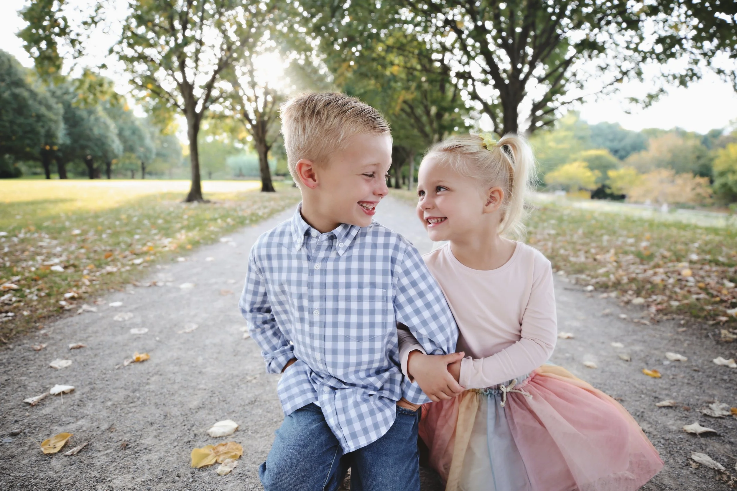 A boy and girl are sitting on a park path, smiling and holding hands. They are surrounded by trees with green and yellow leaves, indicating fall weather.