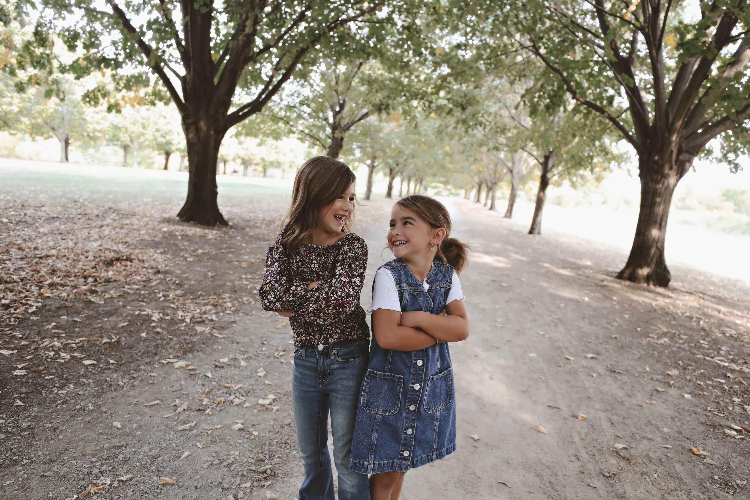 Two young girls with light skin and brown hair smiling and talking in a park with trees and dirt path.