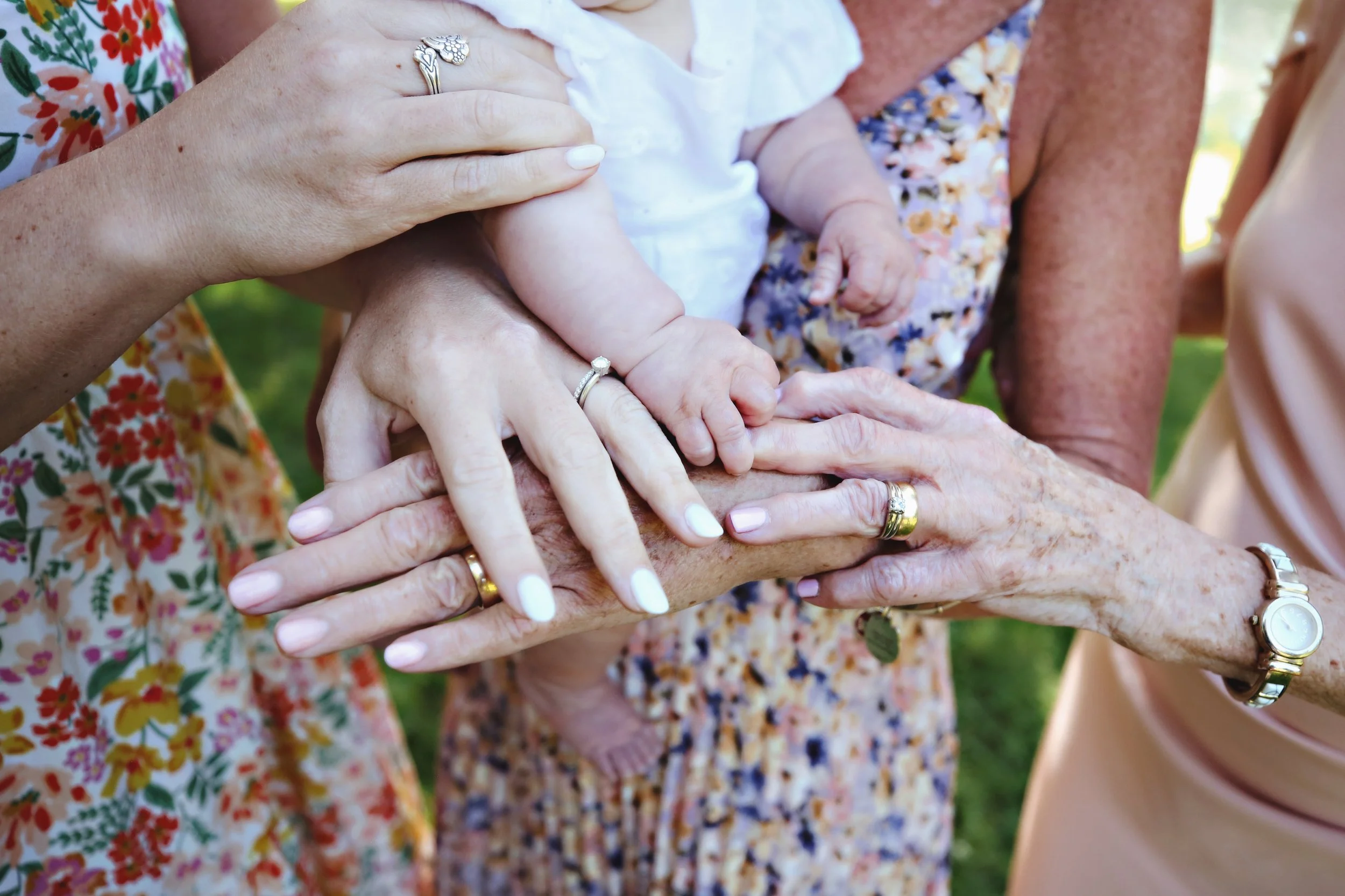 Close-up of multiple hands, including a baby's, touching and holding each other, with visible wedding rings and jewelry, against a background of grass.