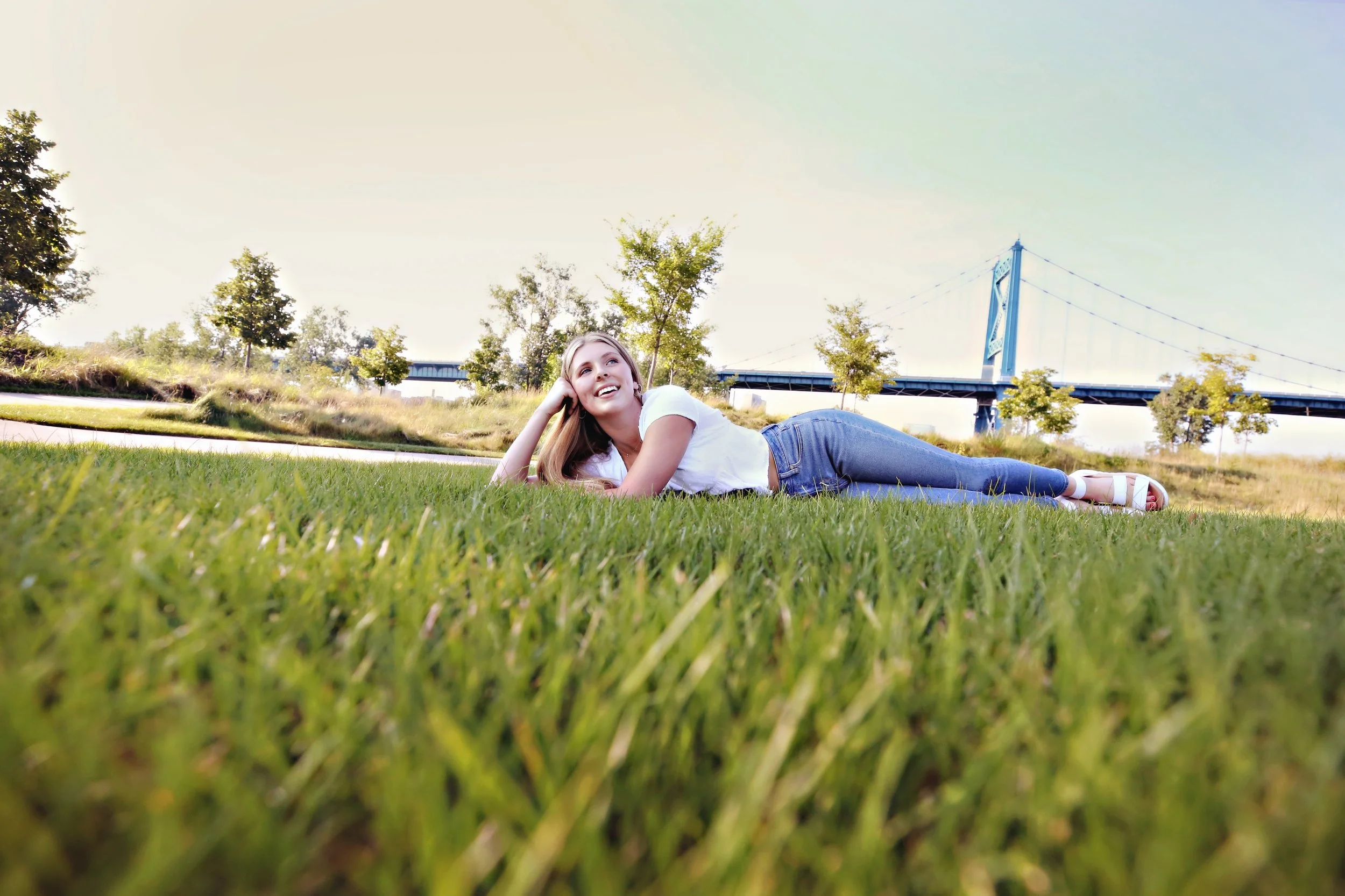 Young woman lying on the grass in a park with trees and a bridge in the background.