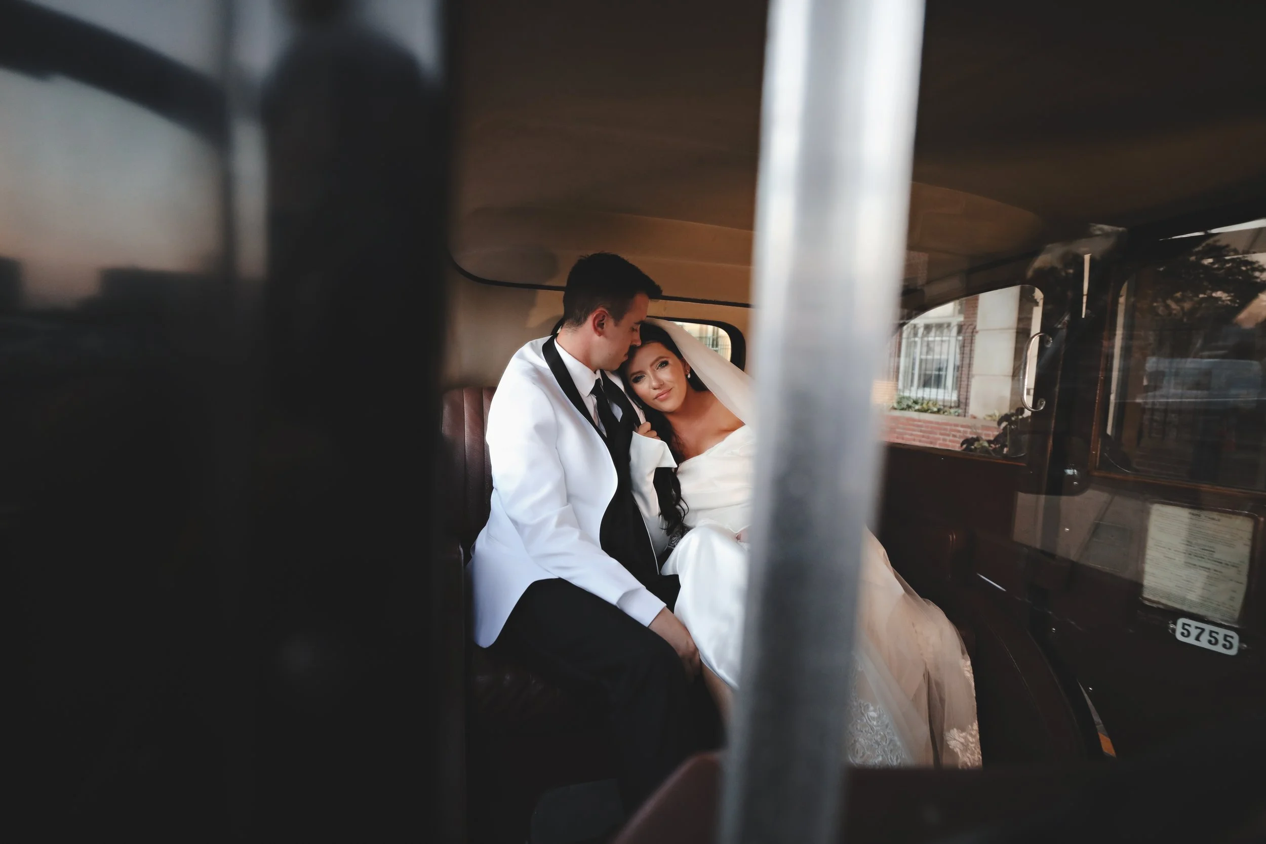 A bride and groom sitting inside a vintage car, with the groom resting his head near the bride as she leans her head on his shoulder, wearing wedding attire.