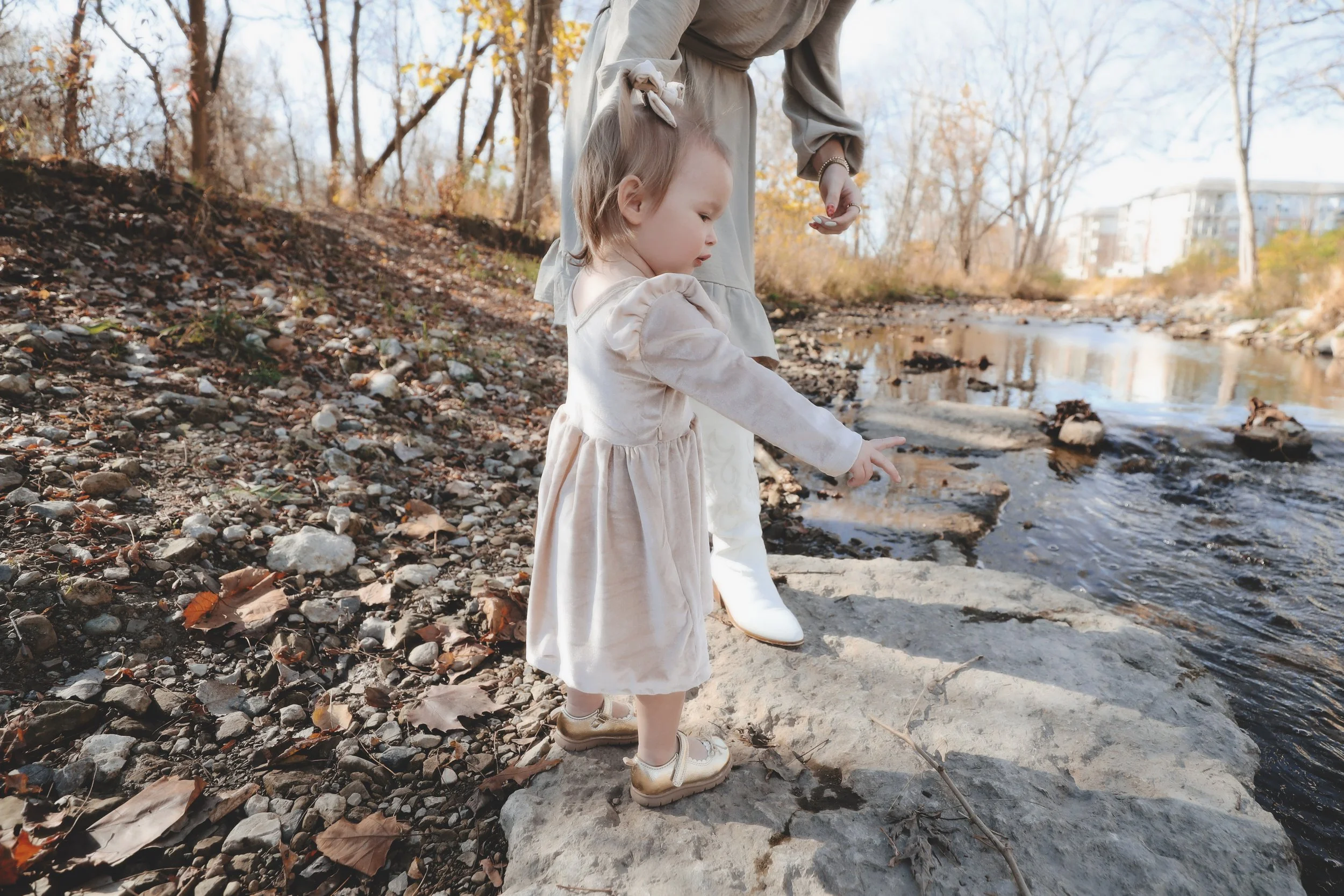 A young girl and an adult, possibly her mother, standing on rocks by a creek pointing at the water. The girl is dressed in a light-colored dress and shoes, with autumn trees in the background.