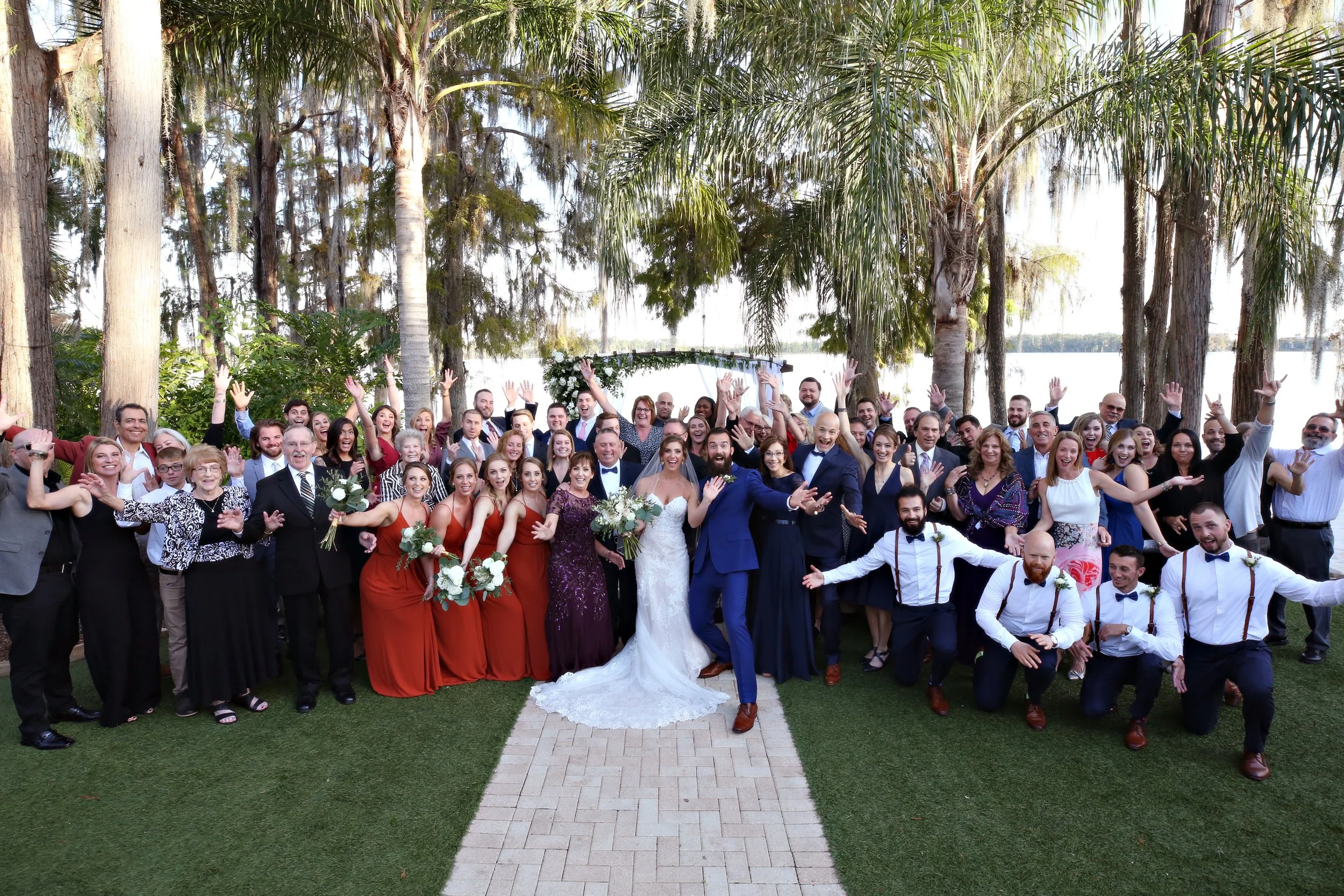 Large group of wedding guests, including bride and groom, posing and celebrating outdoors in front of trees and water, with many raising their hands and smiling.
