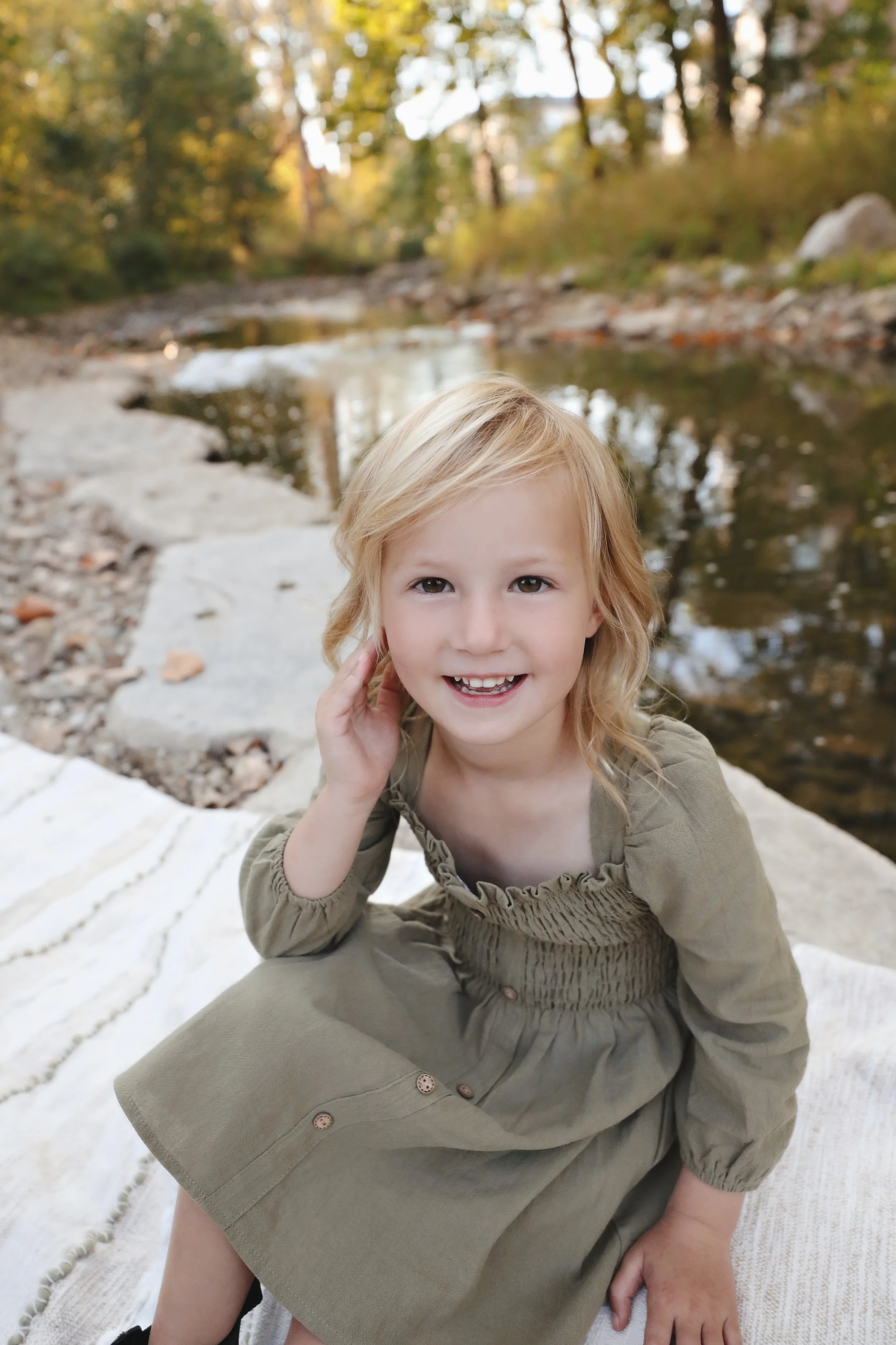 A young girl with blonde hair sitting on a blanket near a small creek surrounded by trees, smiling at the camera.