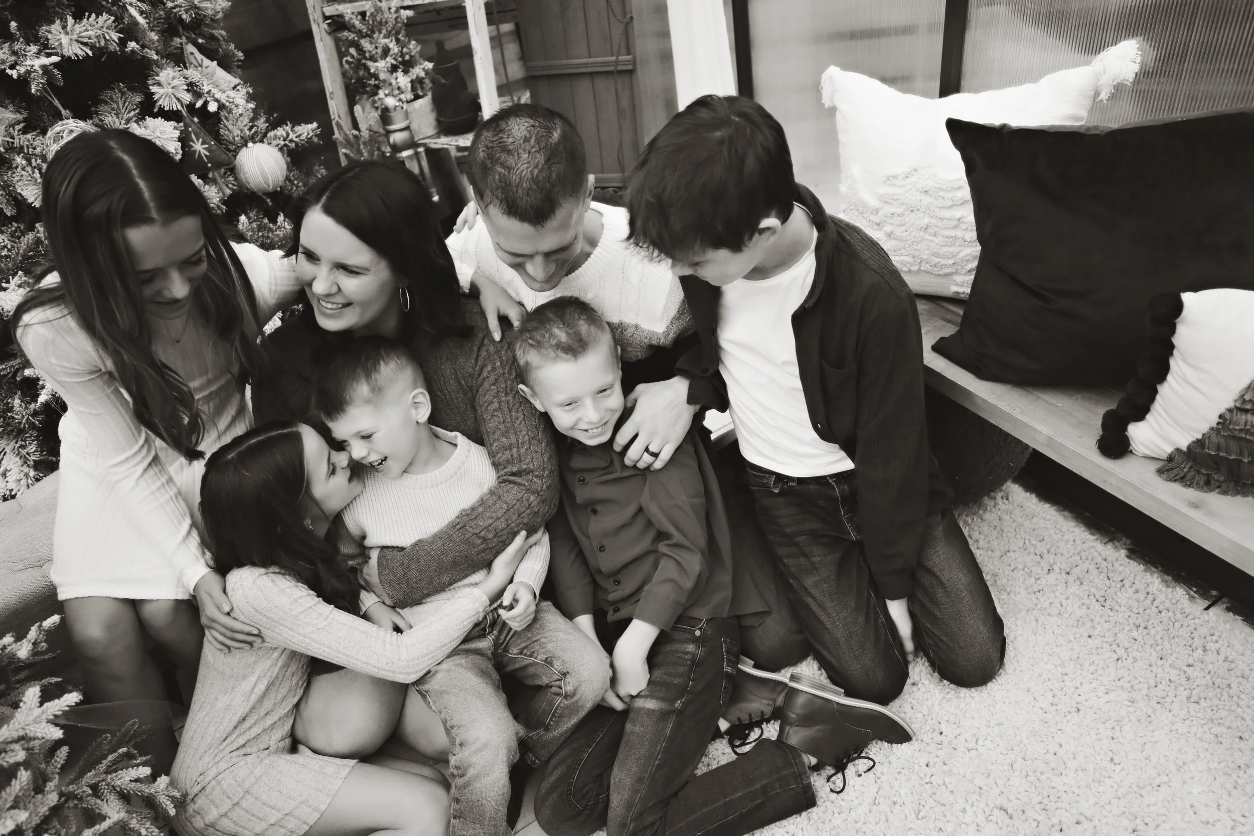 A group of eight children and teenagers gathered together on the floor, smiling and hugging each other, with Christmas decorations and pillows in the background.