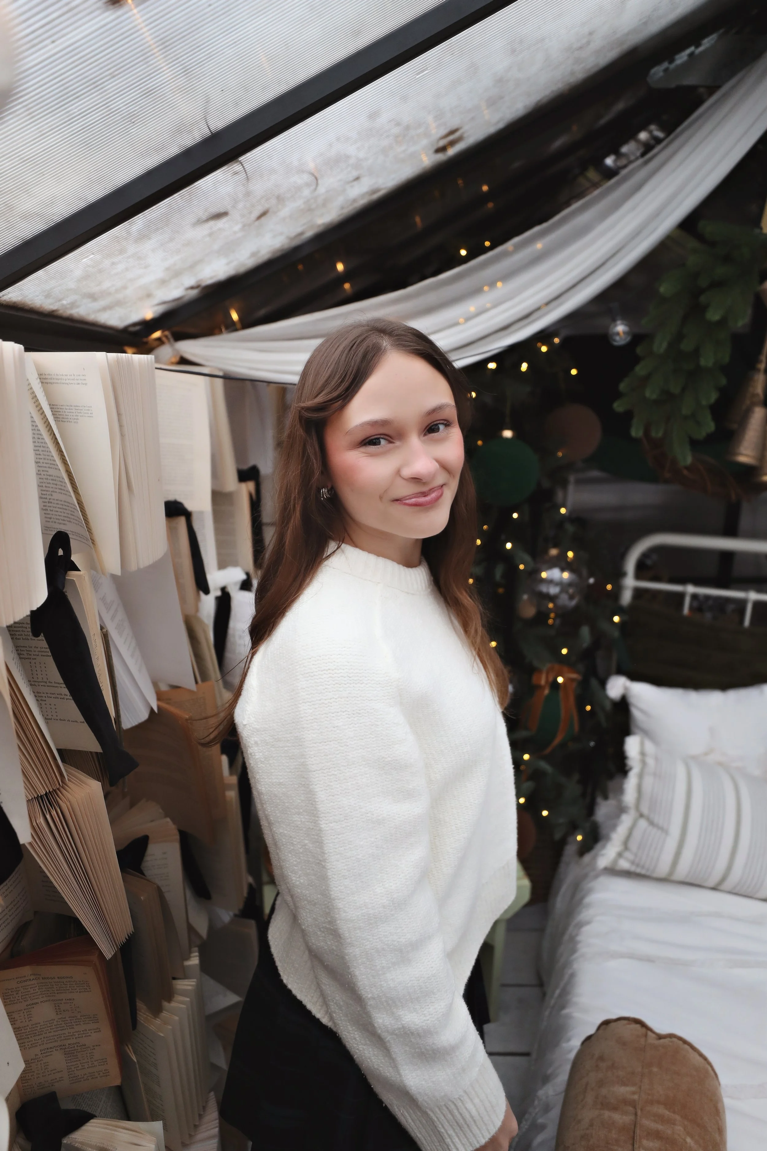 Young woman with long brown hair wearing a white sweater standing indoors near books and a decorated Christmas tree.