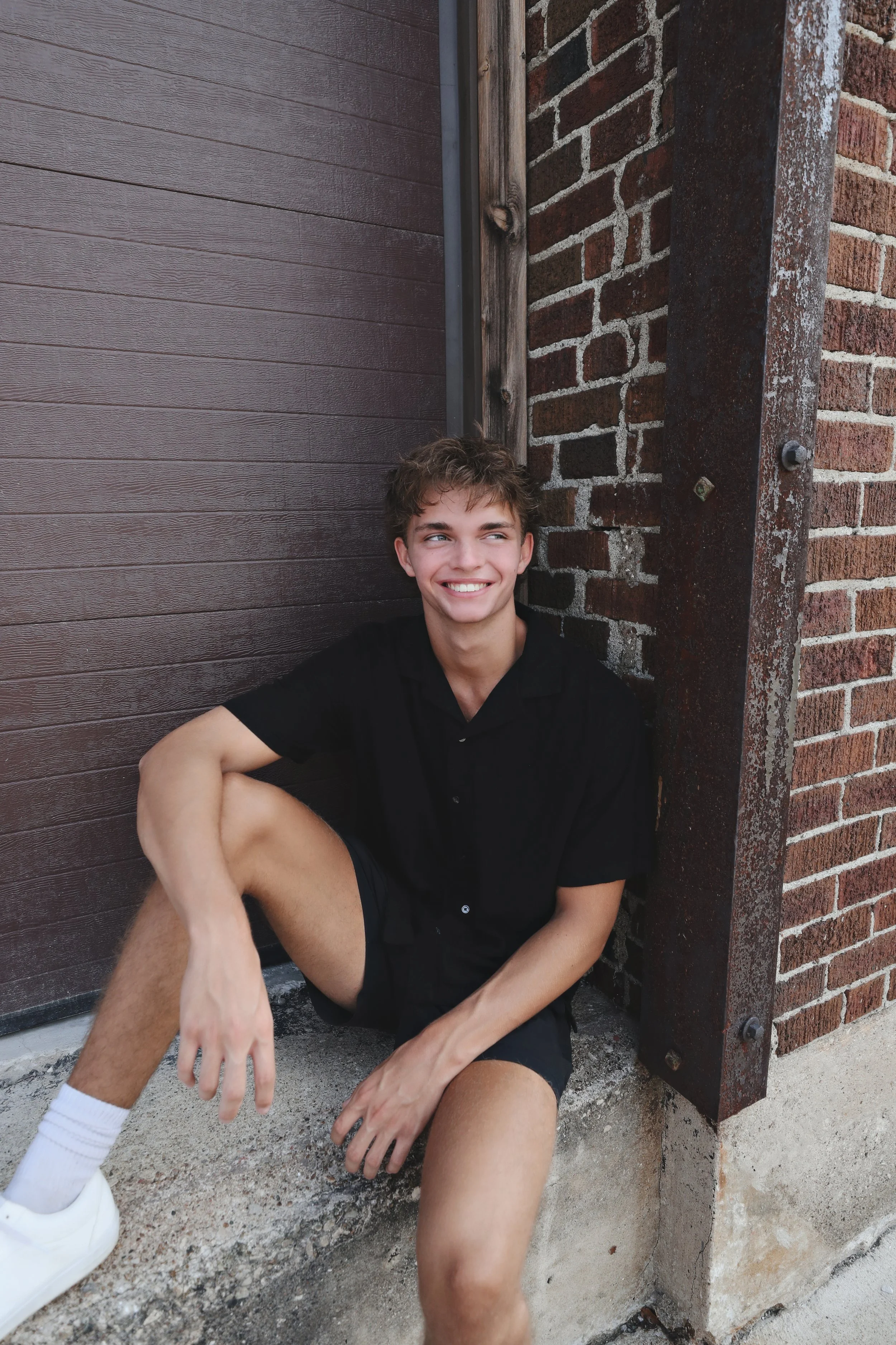 A young man smiling and sitting on the ground, leaning against a brick and wood wall outside.