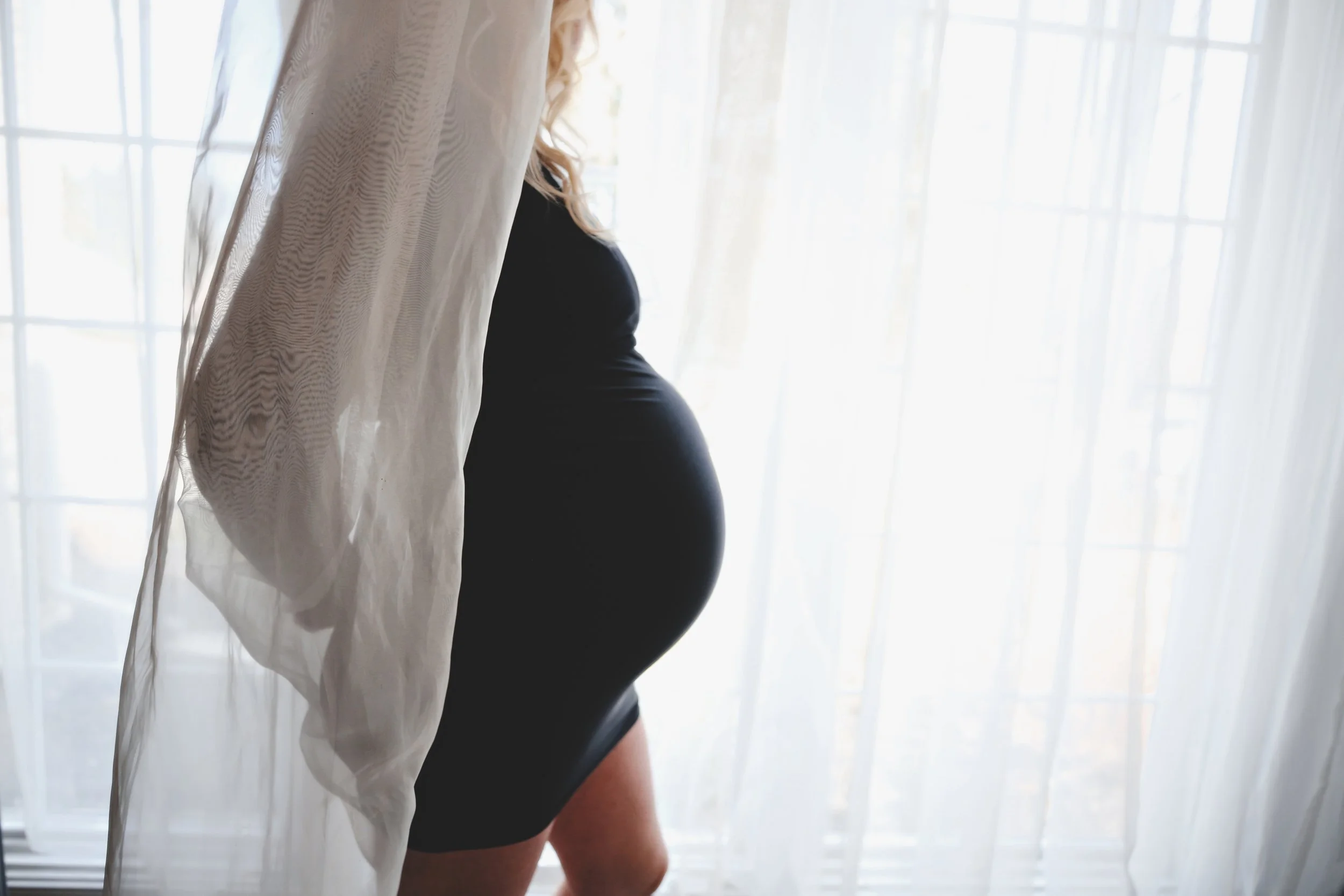 A pregnant woman standing near a window with sheer curtains, wearing a black dress.
