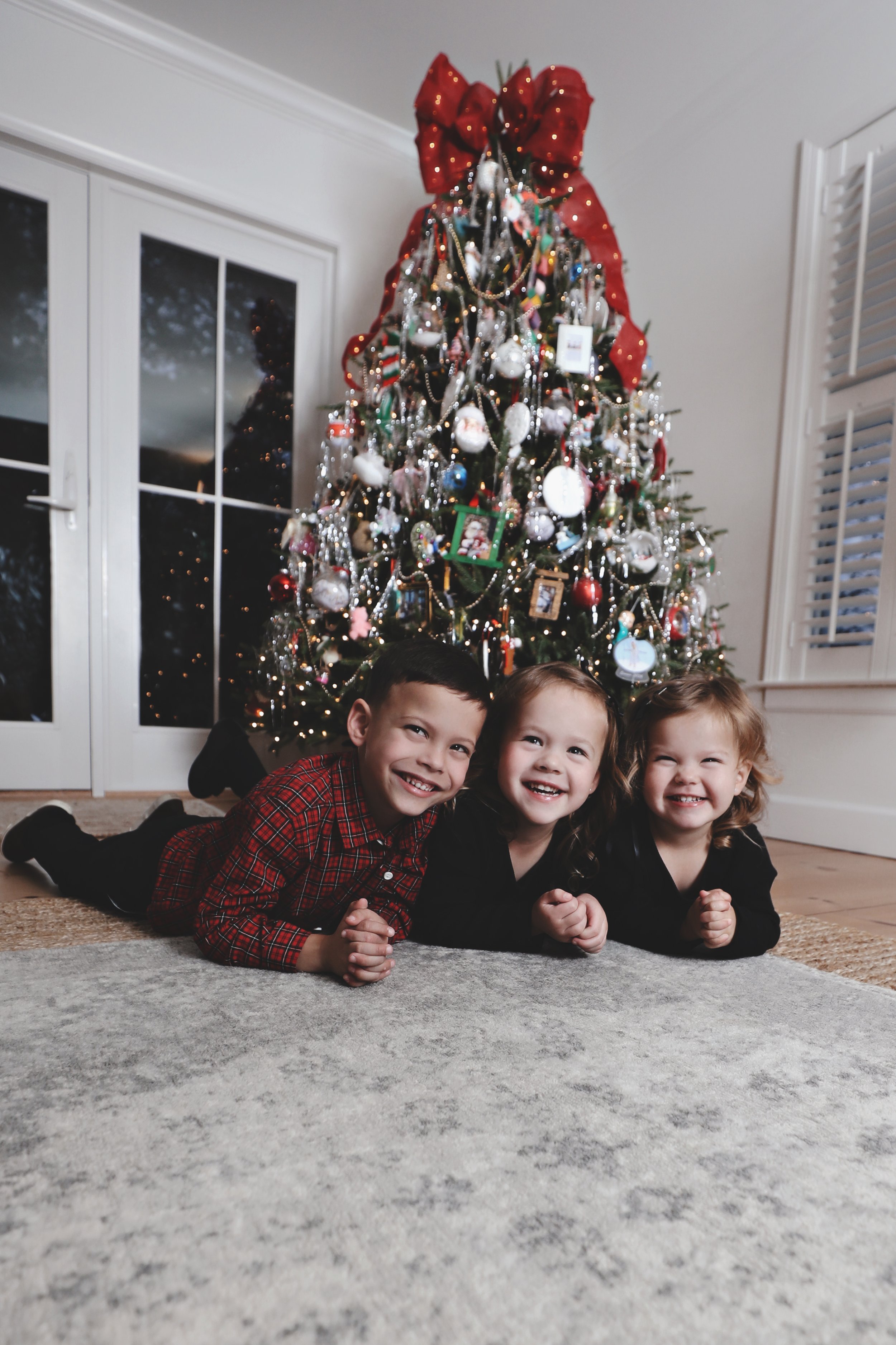 Three children lying on the floor in front of a decorated Christmas tree, smiling.