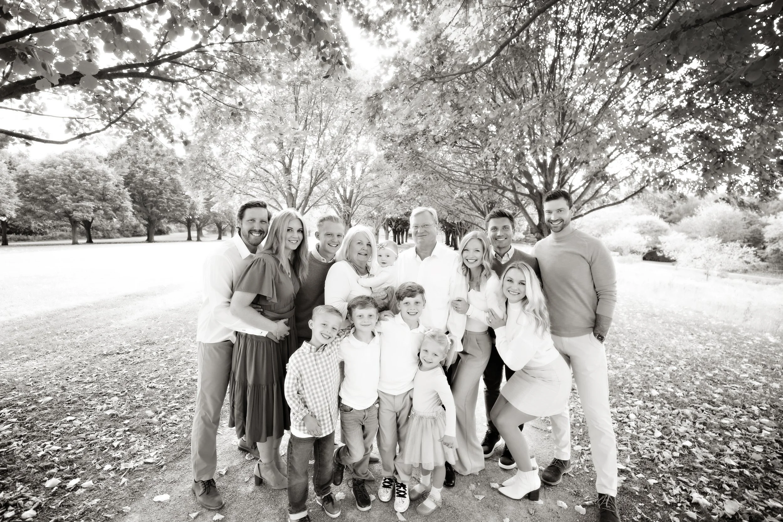 A black and white photo of a large family standing outdoors in a park, with trees and a grassy field in the background. The family members are smiling and posing closely together.