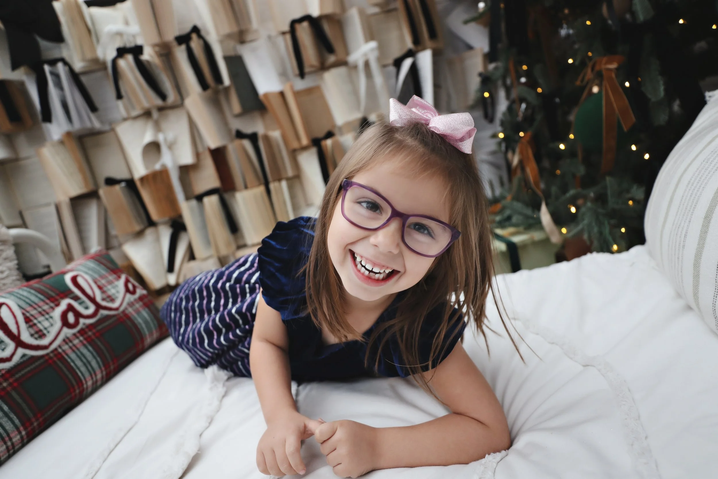 A young girl with glasses and a pink bow on her hair, smiling and lying on her stomach on a white couch, with a Christmas tree and holiday decorations in the background.
