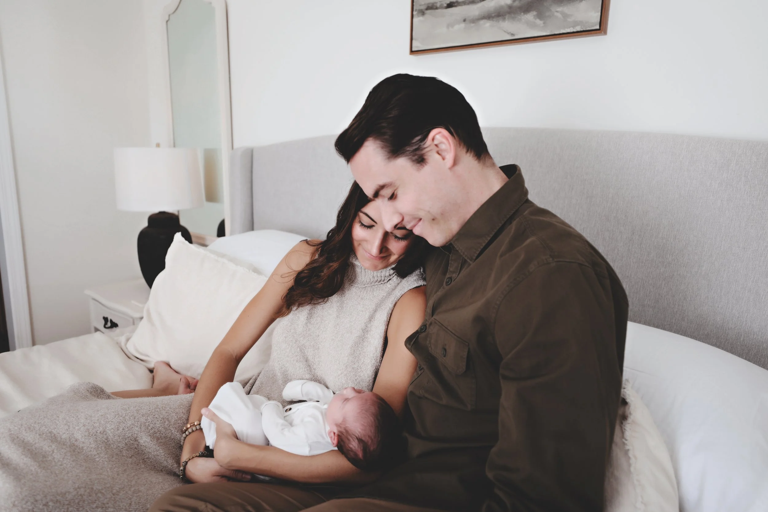 A couple sitting on a bed, holding their newborn baby, smiling and looking at each other affectionately.