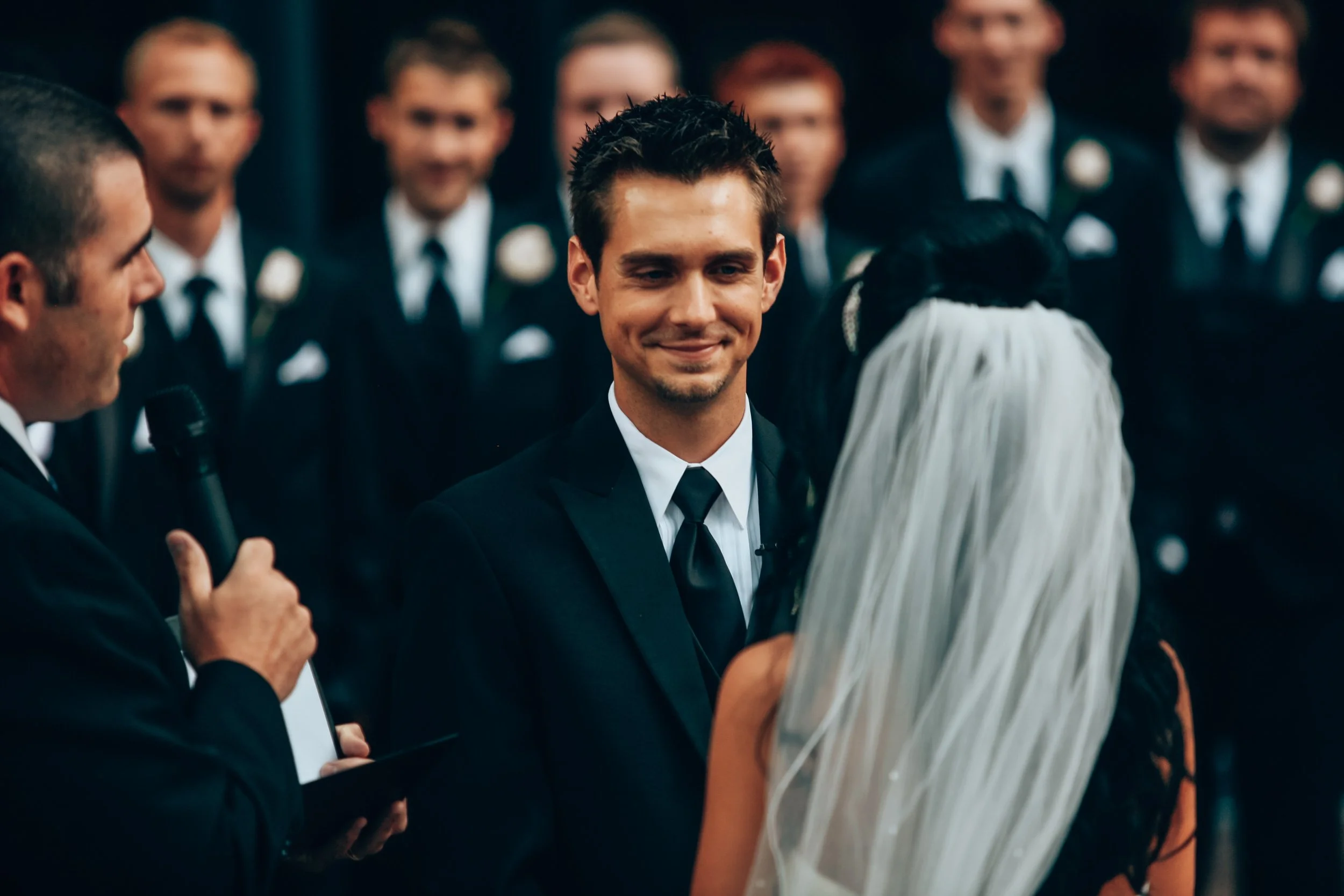 A groom smiling at a bride during a wedding ceremony in front of an officiant with a microphone, with groomsmen in the background.