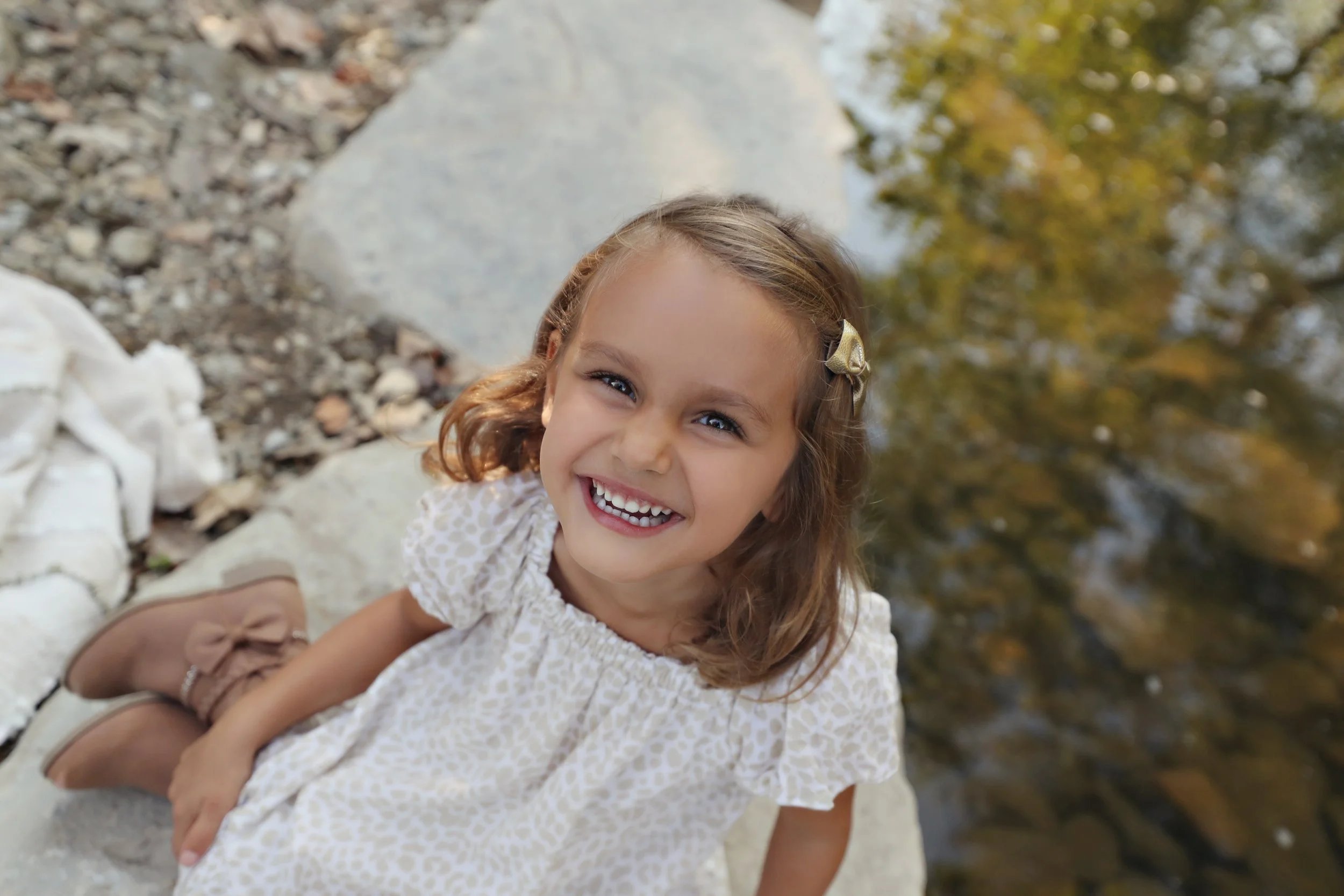 A young girl with curly brown hair, wearing a beige patterned dress and brown shoes, sitting on a rock by a body of water, smiling at the camera.