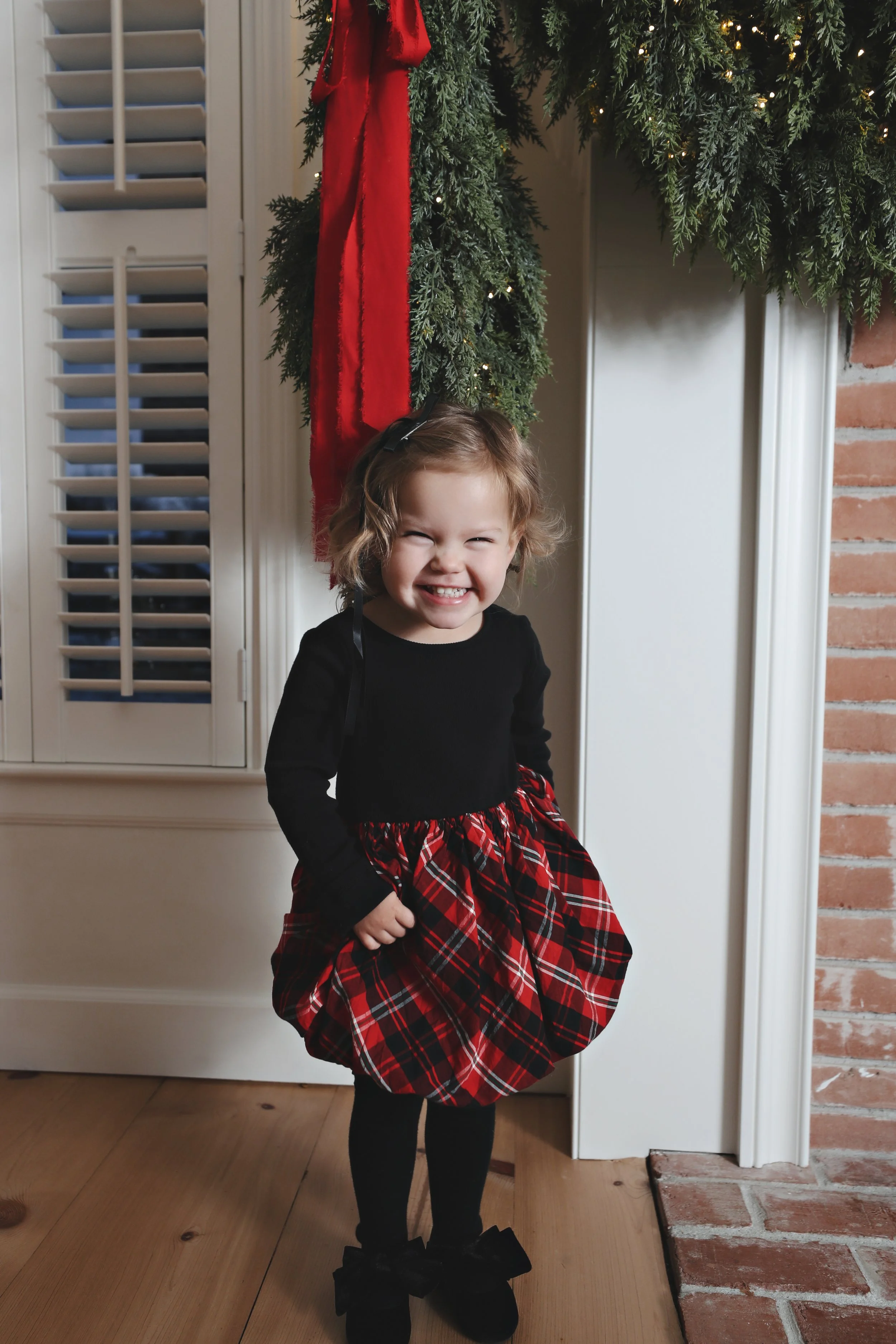 A young girl with curly blonde hair smiling cheerfully in front of a decorated Christmas fireplace with greenery, red ribbon, and lights, wearing a black long-sleeve top and a red plaid dress with black tights and shoes.