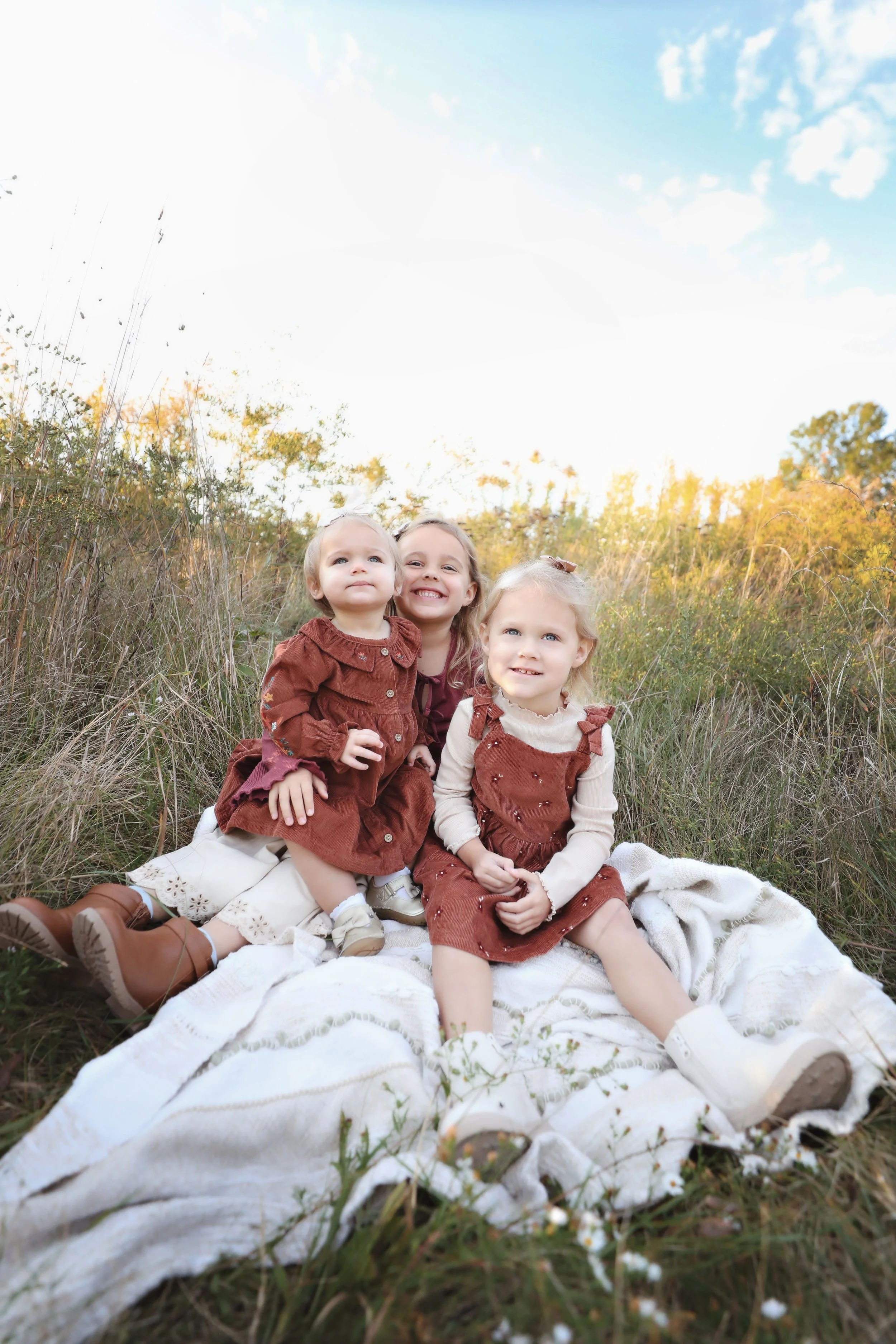 Three young girls sitting on a white blanket in a field of tall grass during autumn, smiling and posing for a photo under a partly cloudy sky.