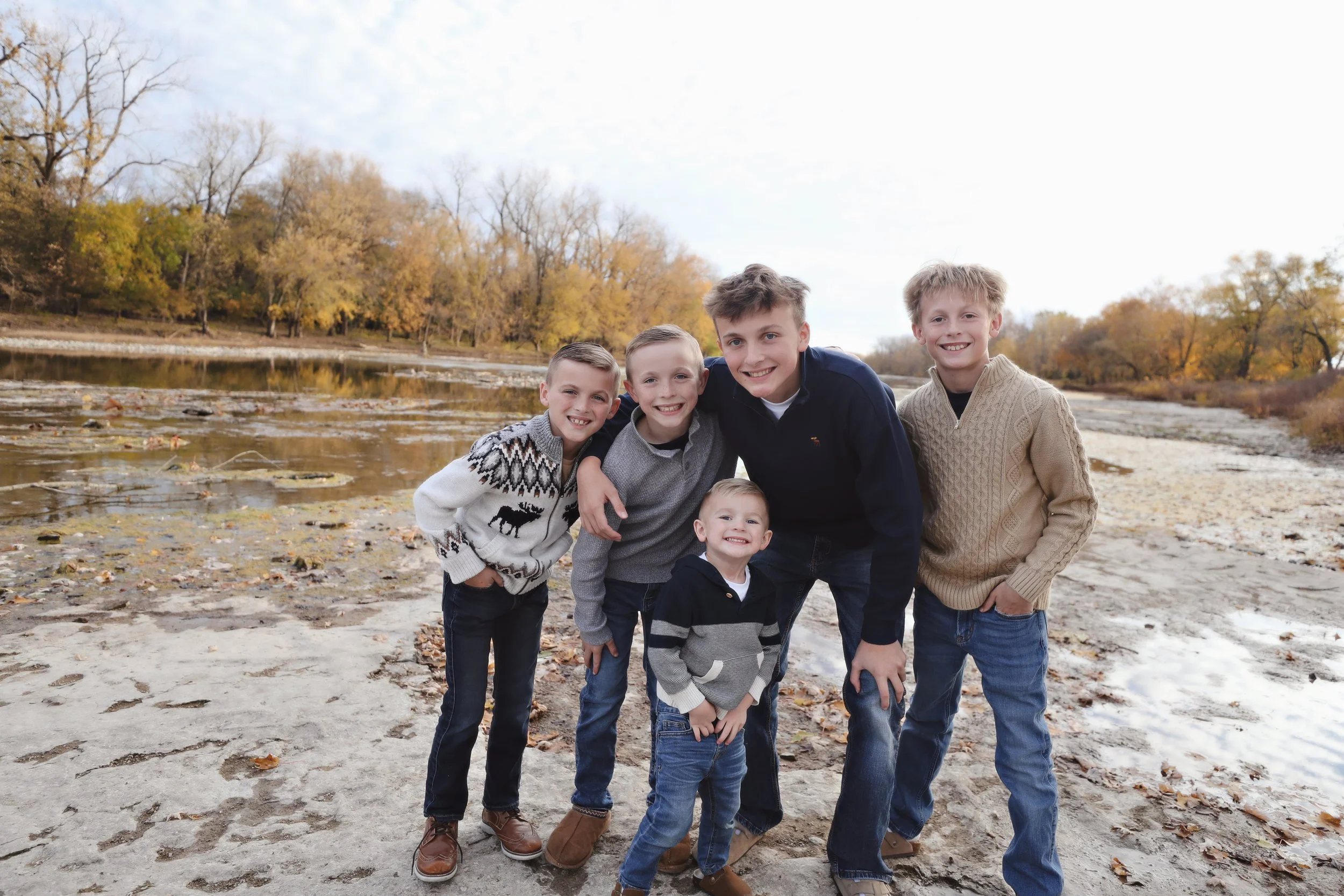 A group of six boys smiling and standing together outdoors near a river during fall, with trees in the background.
