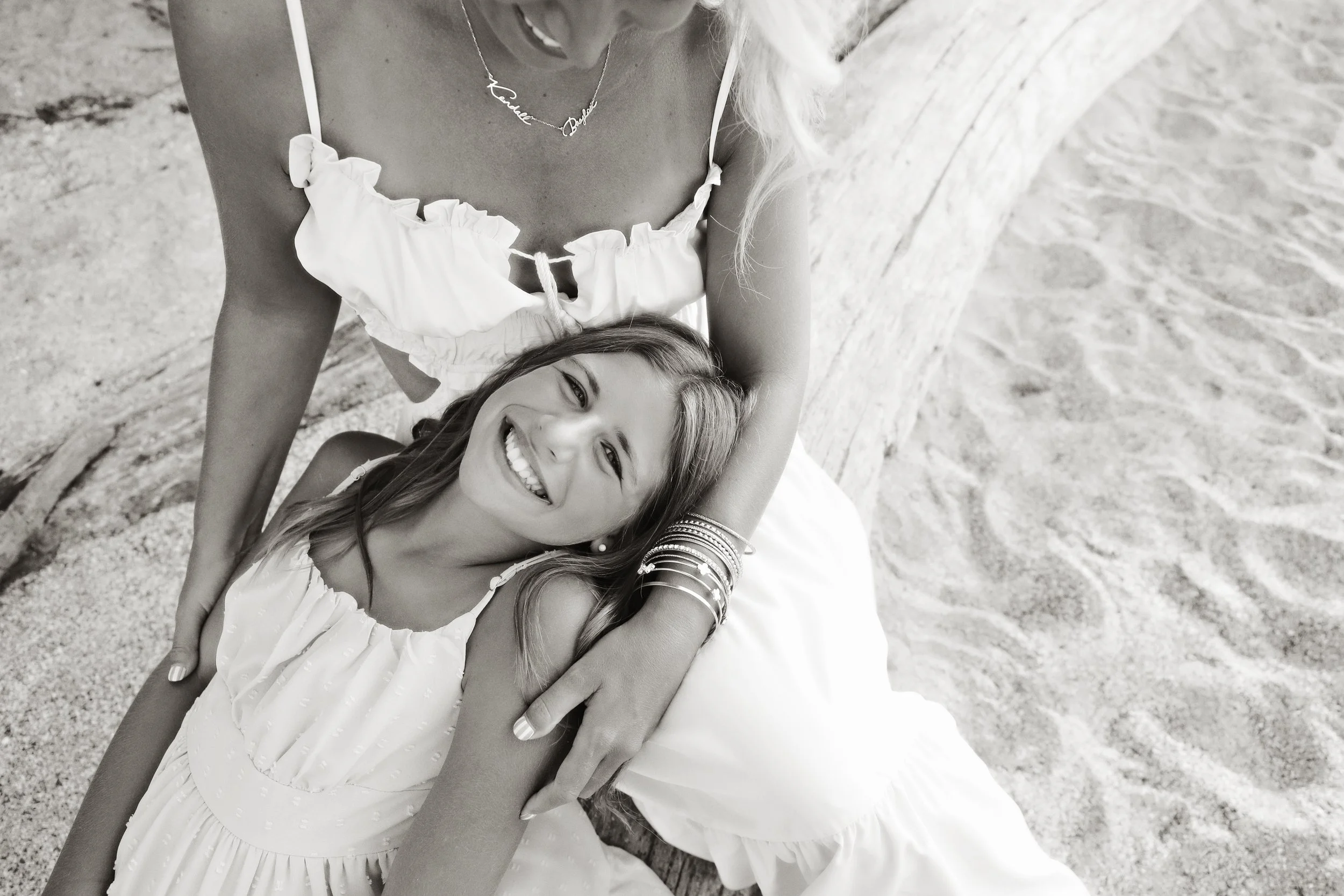 A young woman with long hair smiling while lying on the sand, with an older woman partially visible beside her, both wearing white dresses. They are at the beach, and the older woman has her arm around the young woman.
