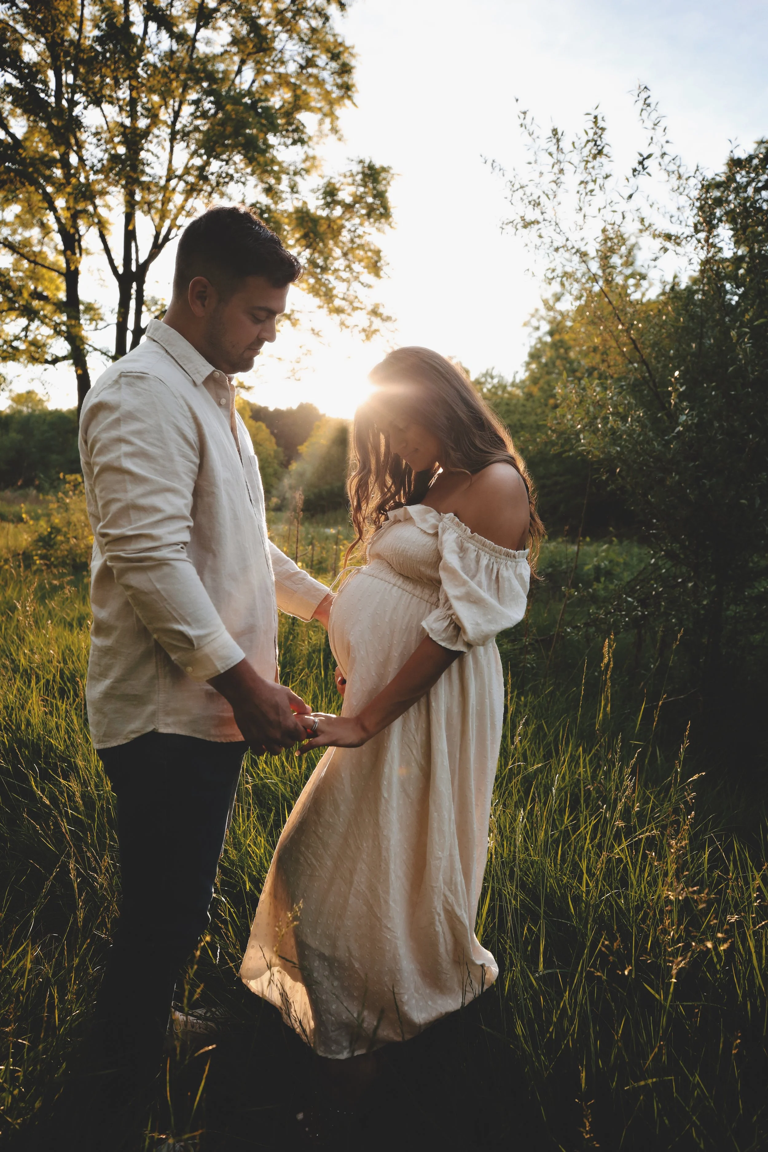 A pregnant woman in a white dress holds hands with a man in a white shirt in a grassy outdoor setting during sunset.
