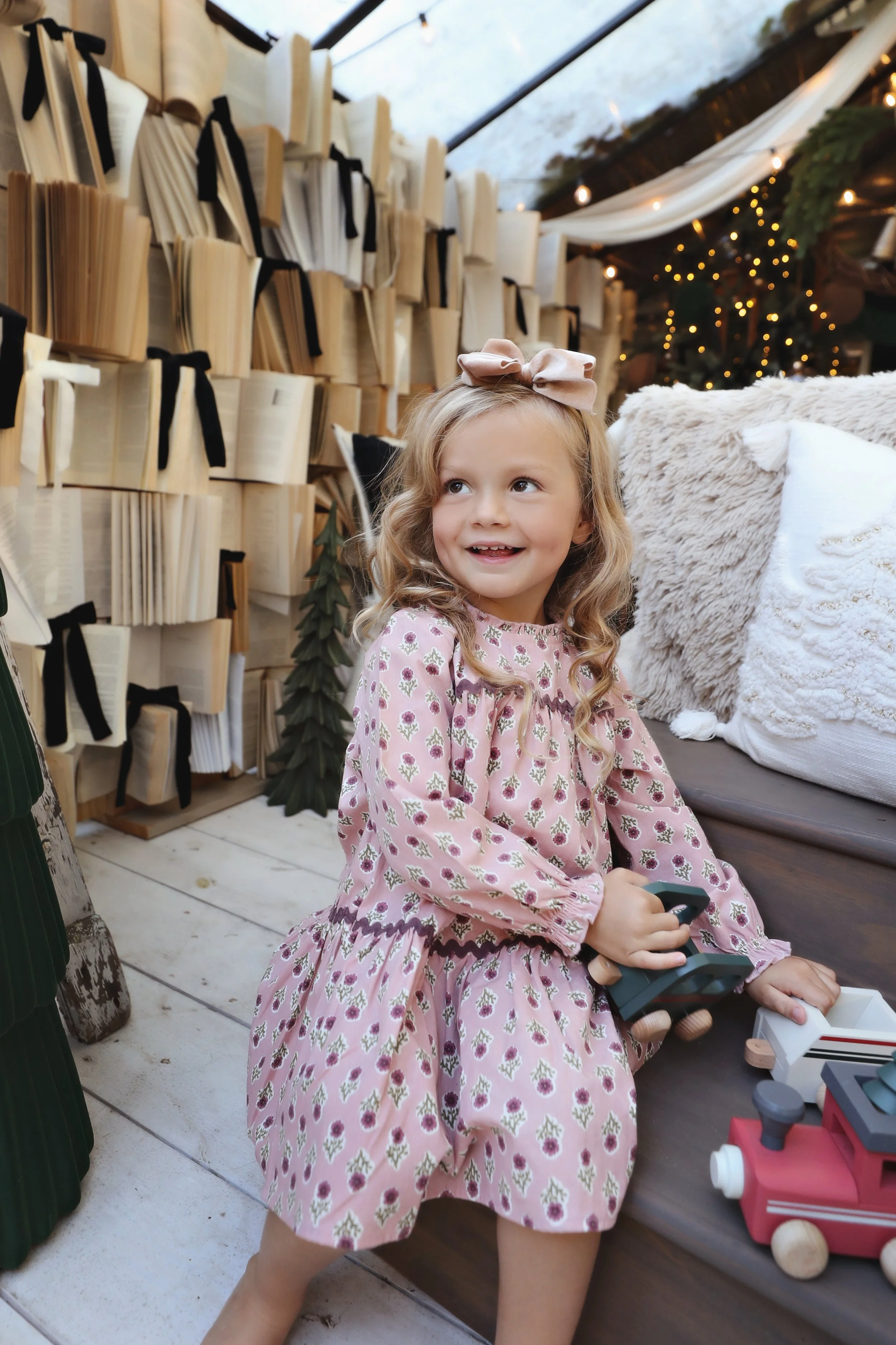 A young girl with long curly blonde hair and a pink bow in her hair, sitting on a wooden bench, smiling and playing with toy trains inside a cozy, decorated space with books and string lights.