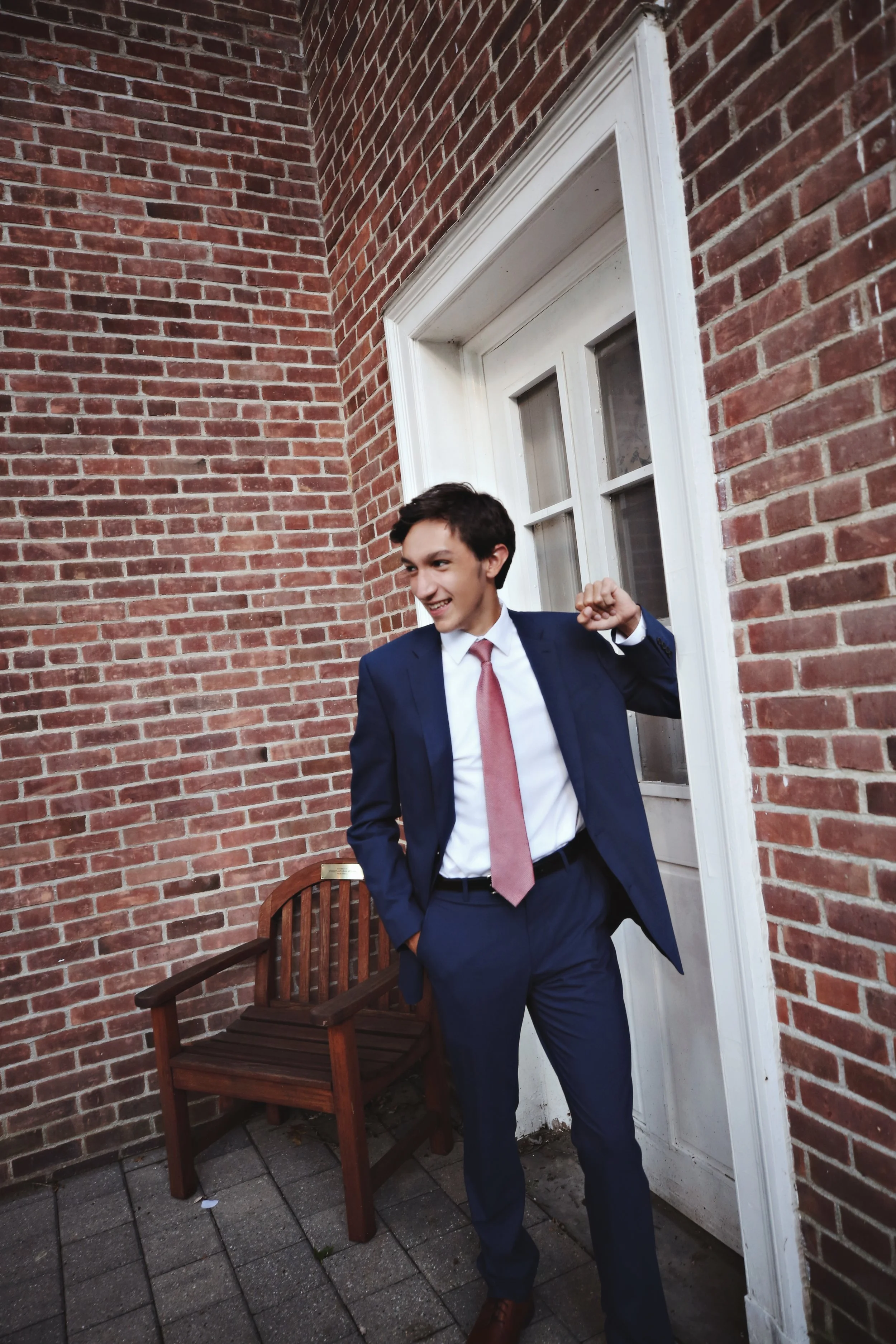A young man in a navy suit, white shirt, and pink tie standing outside near a white door, smiling and making a fist pump gesture.