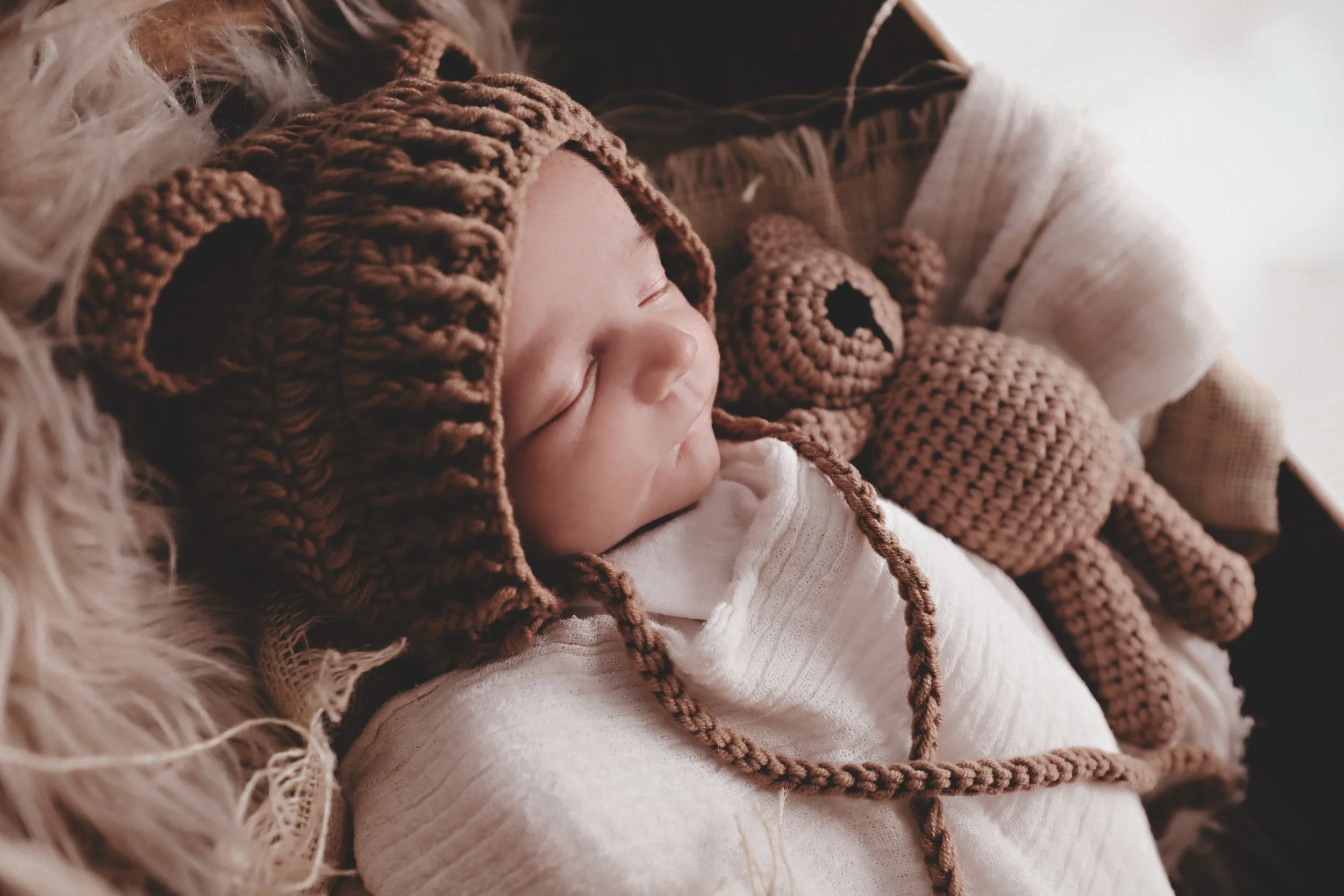 A sleeping baby wearing a brown crocheted bear hat with bear ears, lying on a fluffy blanket, holding a crocheted bear stuffed toy.