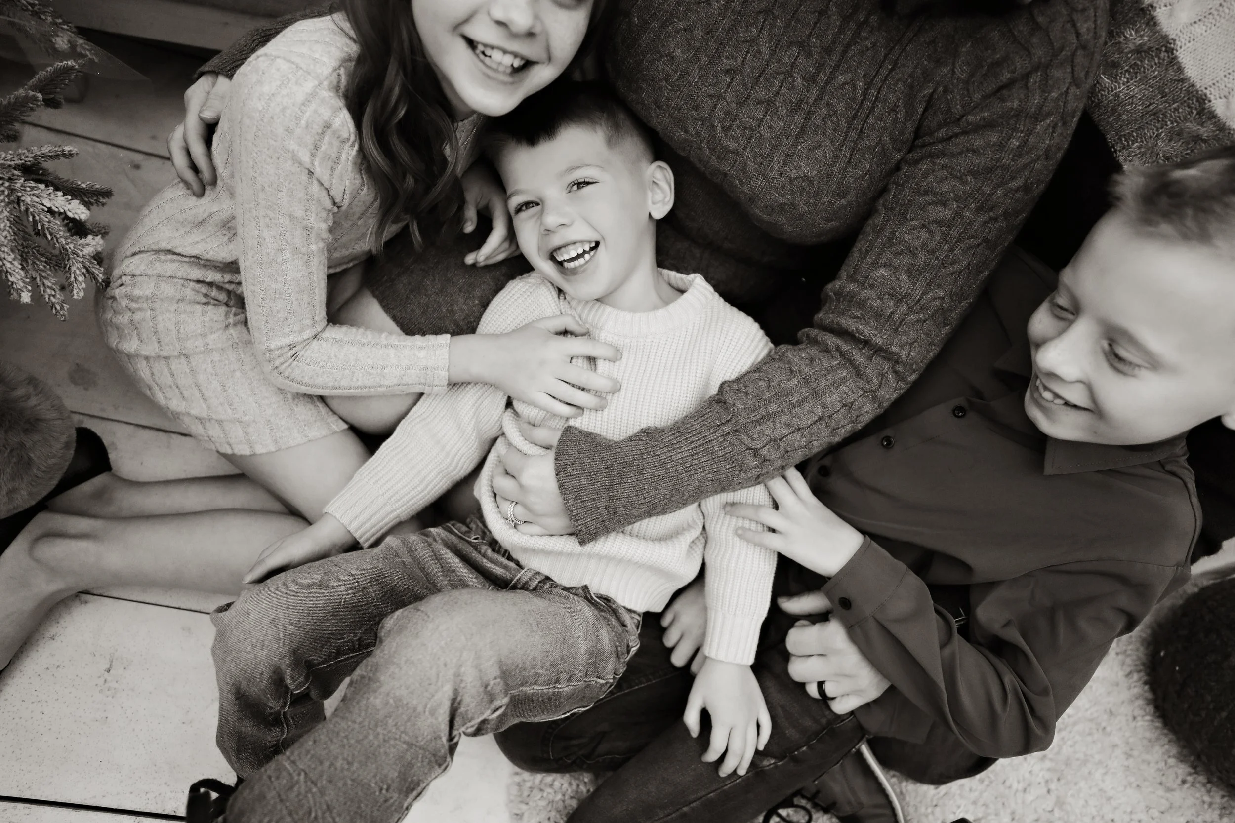 Three children, two boys and a girl, are playfully lying together on the floor, smiling and laughing. One girl with long hair is on the left, a boy with short hair in the middle, and another boy with short hair on the right. They are interacting clos