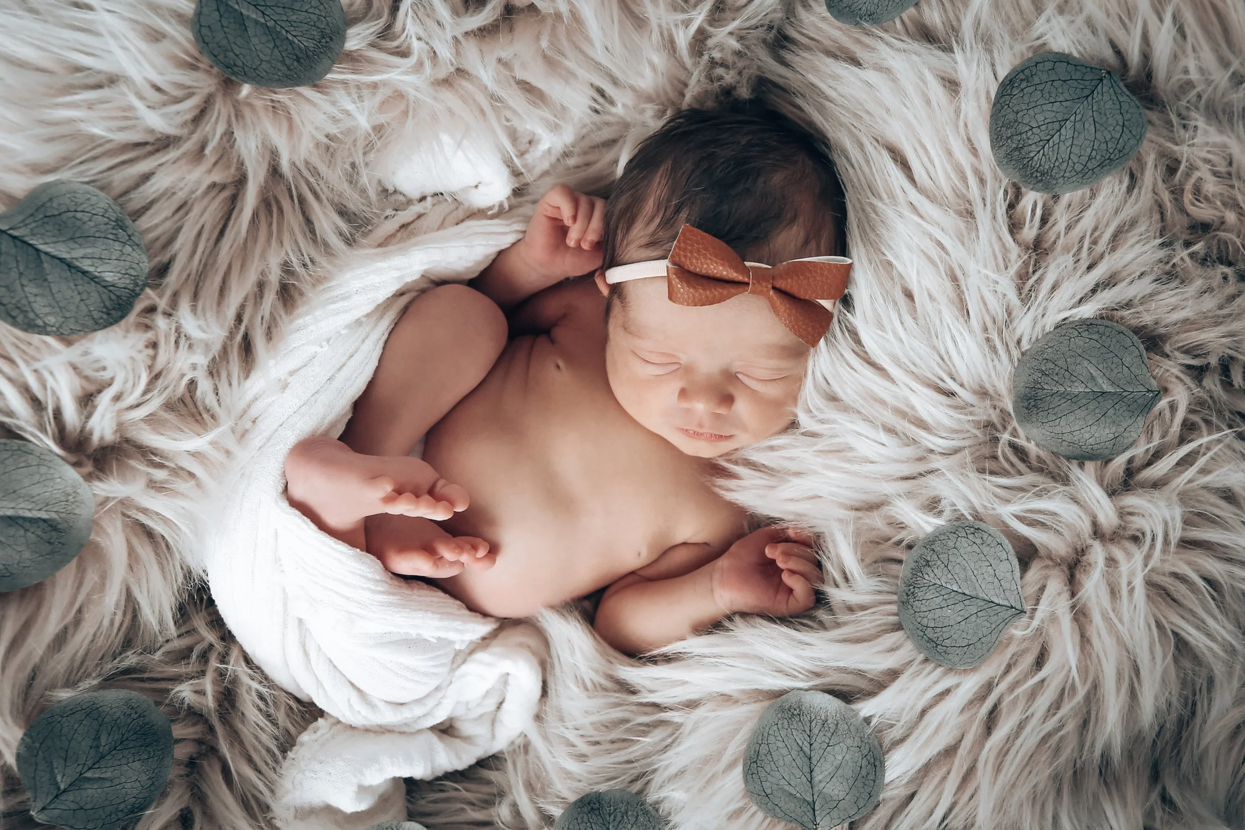 A newborn baby girl sleeping peacefully on a fluffy cream-colored blanket, wearing a small brown bow headband and wrapped in a white cloth, surrounded by green leaves.