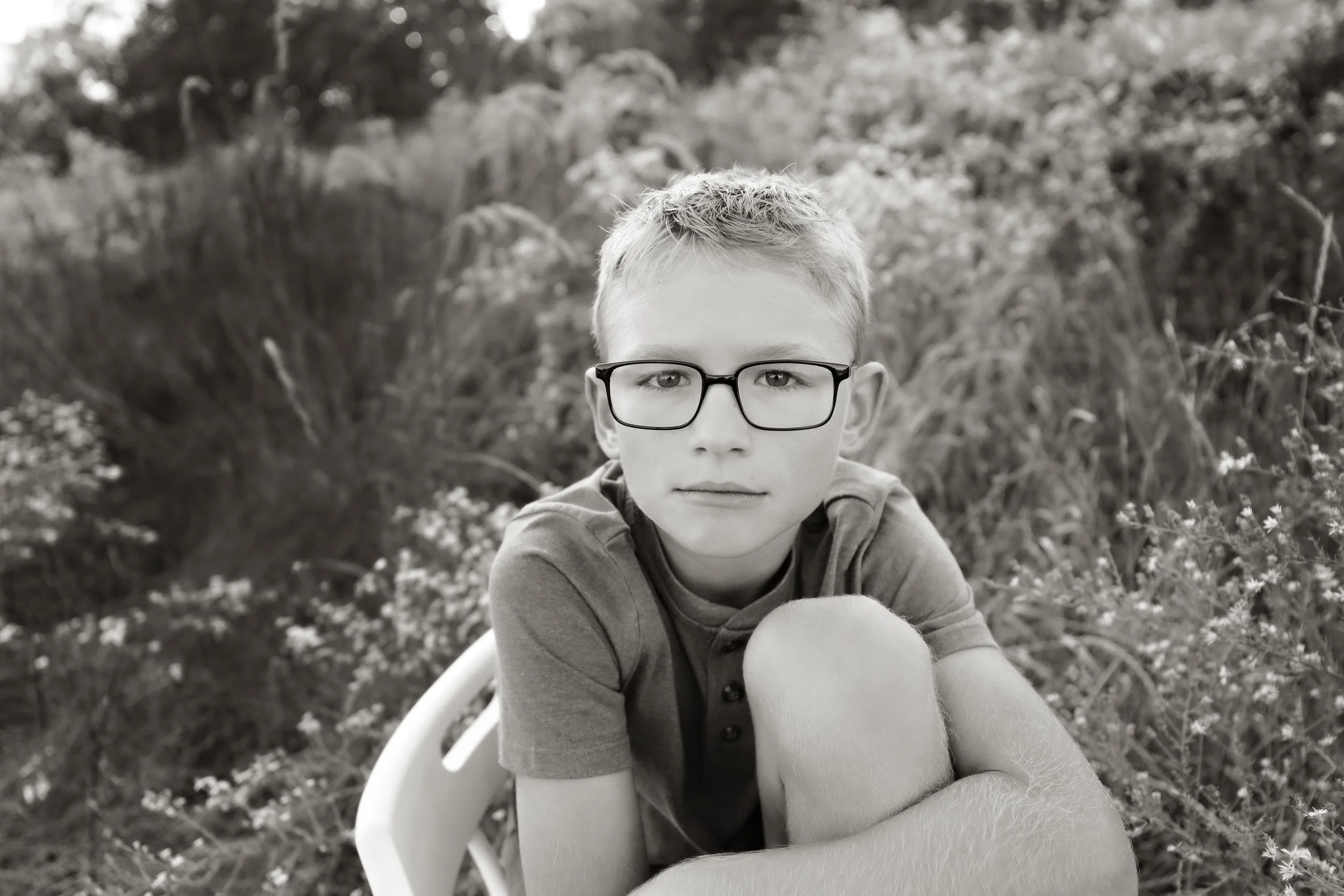 A black-and-white photo of a young boy with glasses sitting outdoors among flowers and plants, looking directly at the camera.