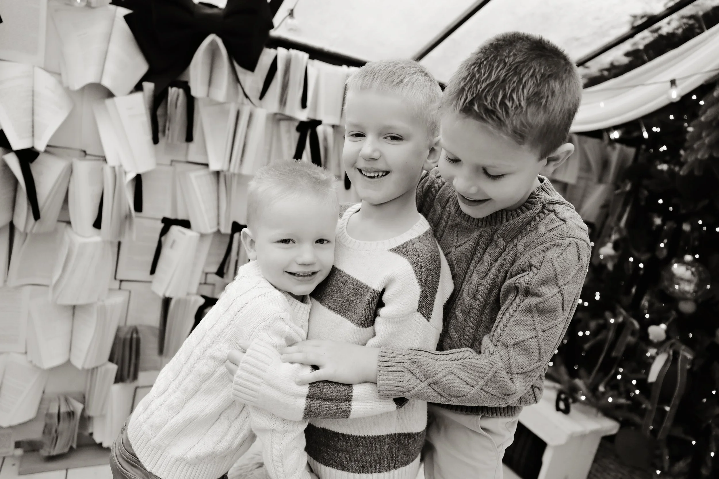 Three young boys embracing each other in front of a cozy Christmas or holiday display, with books and lights in the background.