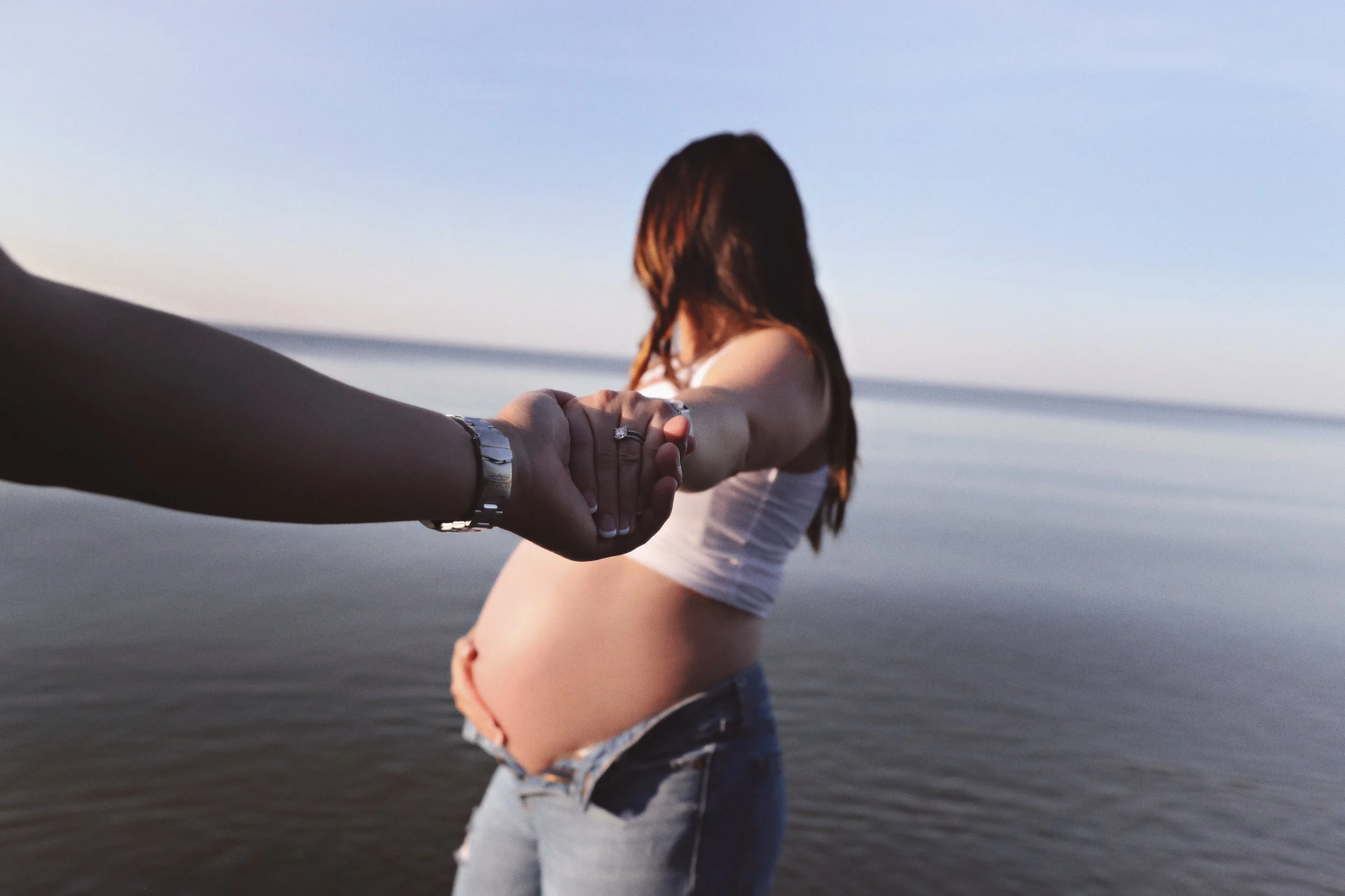 A pregnant woman holding hands with another person on the beach at sunset, with the ocean in the background.