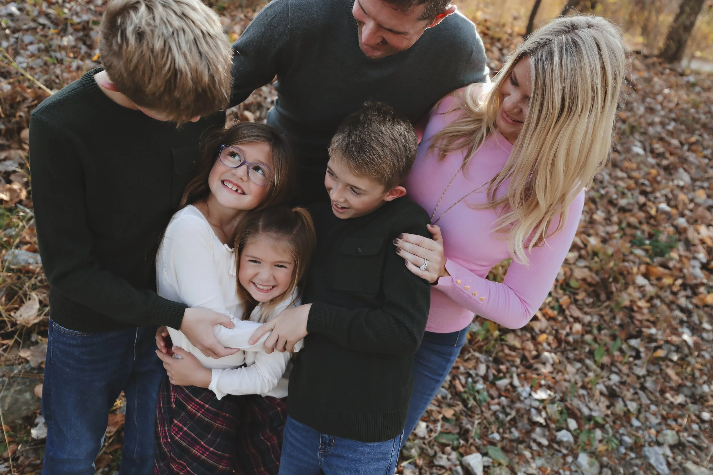A family of six, including two teenagers, a young girl, a young boy, and an adult woman and man, hugging and smiling outdoors during fall with fallen leaves on the ground.