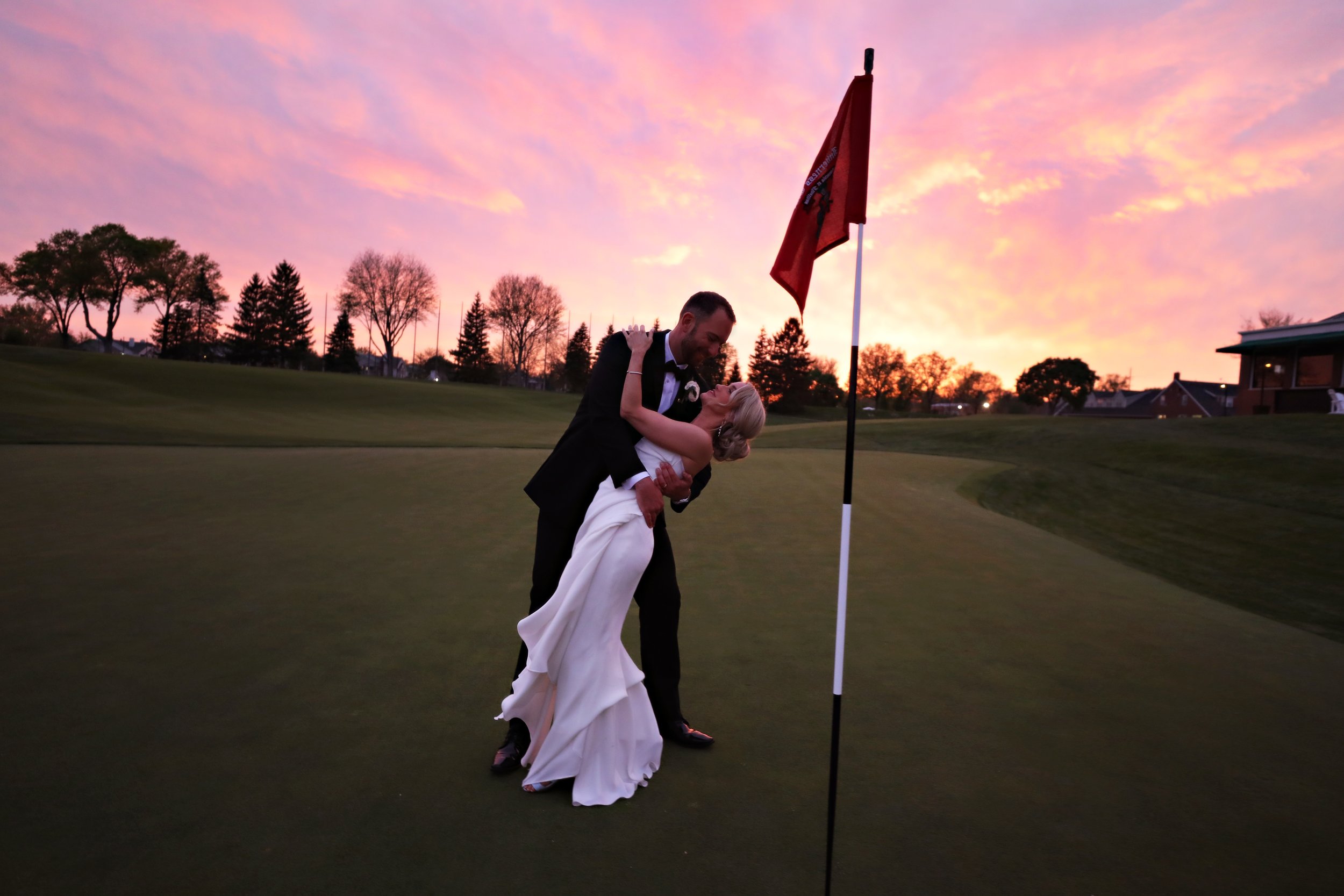 A newlywed couple in wedding attire on a golf course at sunset, with the groom dipping the bride and they are close to a flagstick.