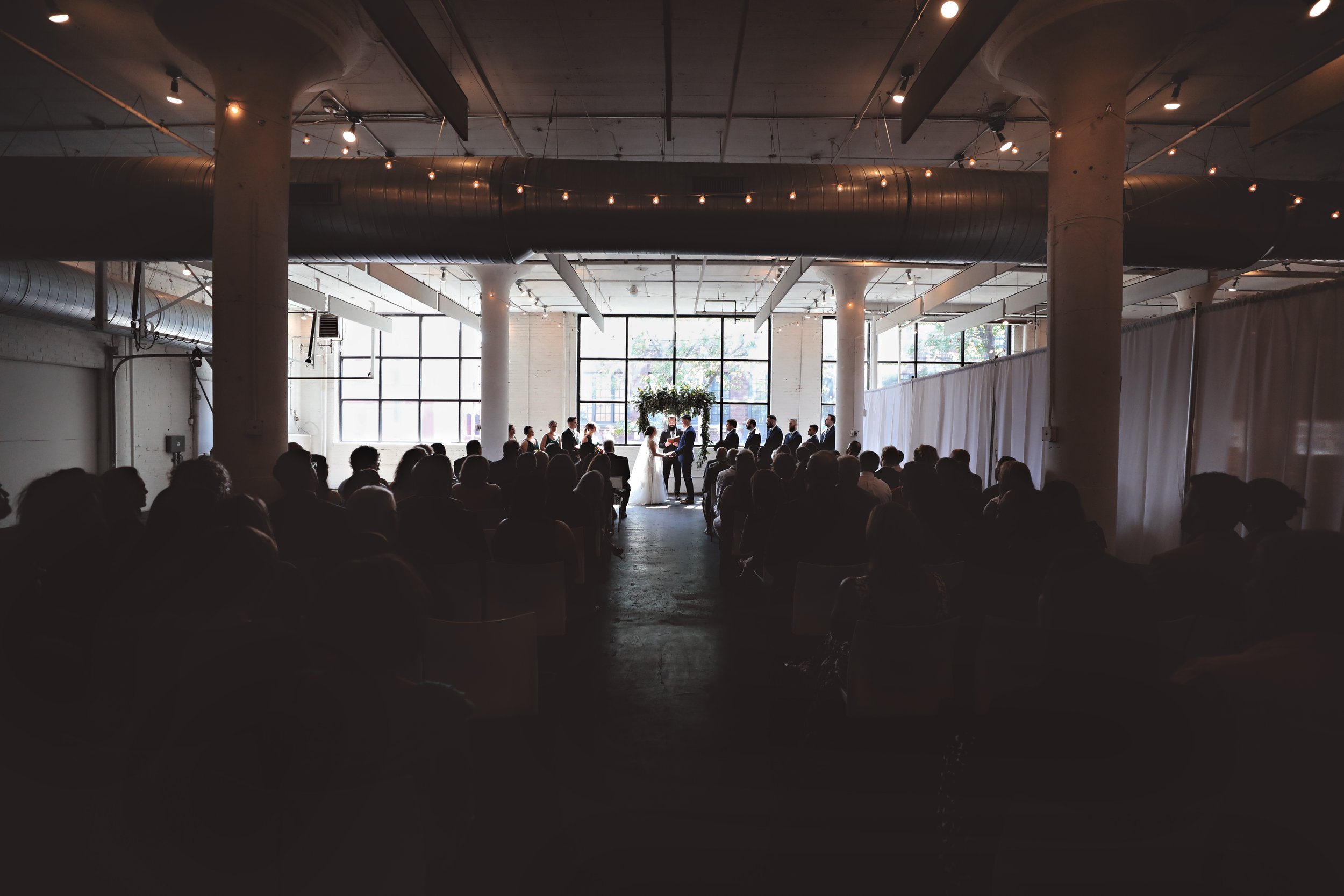 Indoor wedding ceremony with a bride and groom at the altar, surrounded by guests sitting in rows. The setting is bright with large windows, industrial-style decor, and string lights overhead.