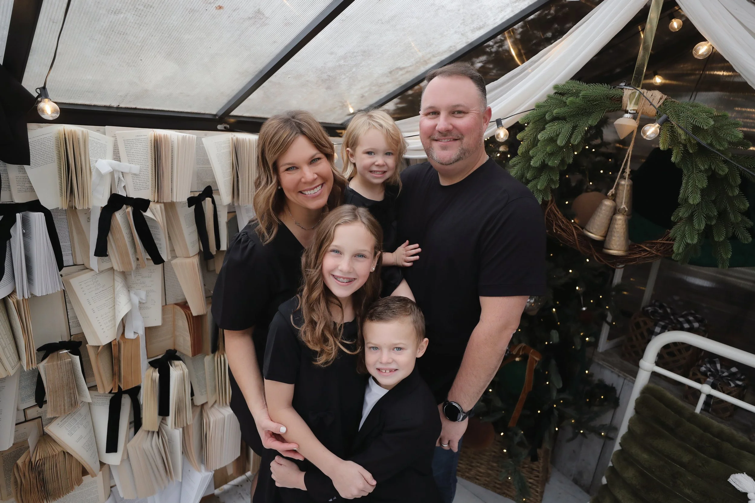 A family of six smiling, wearing black shirts, standing inside a decorated area with open books tied with black ribbons on the wall, and a Christmas tree with lights and gold bells in the background.