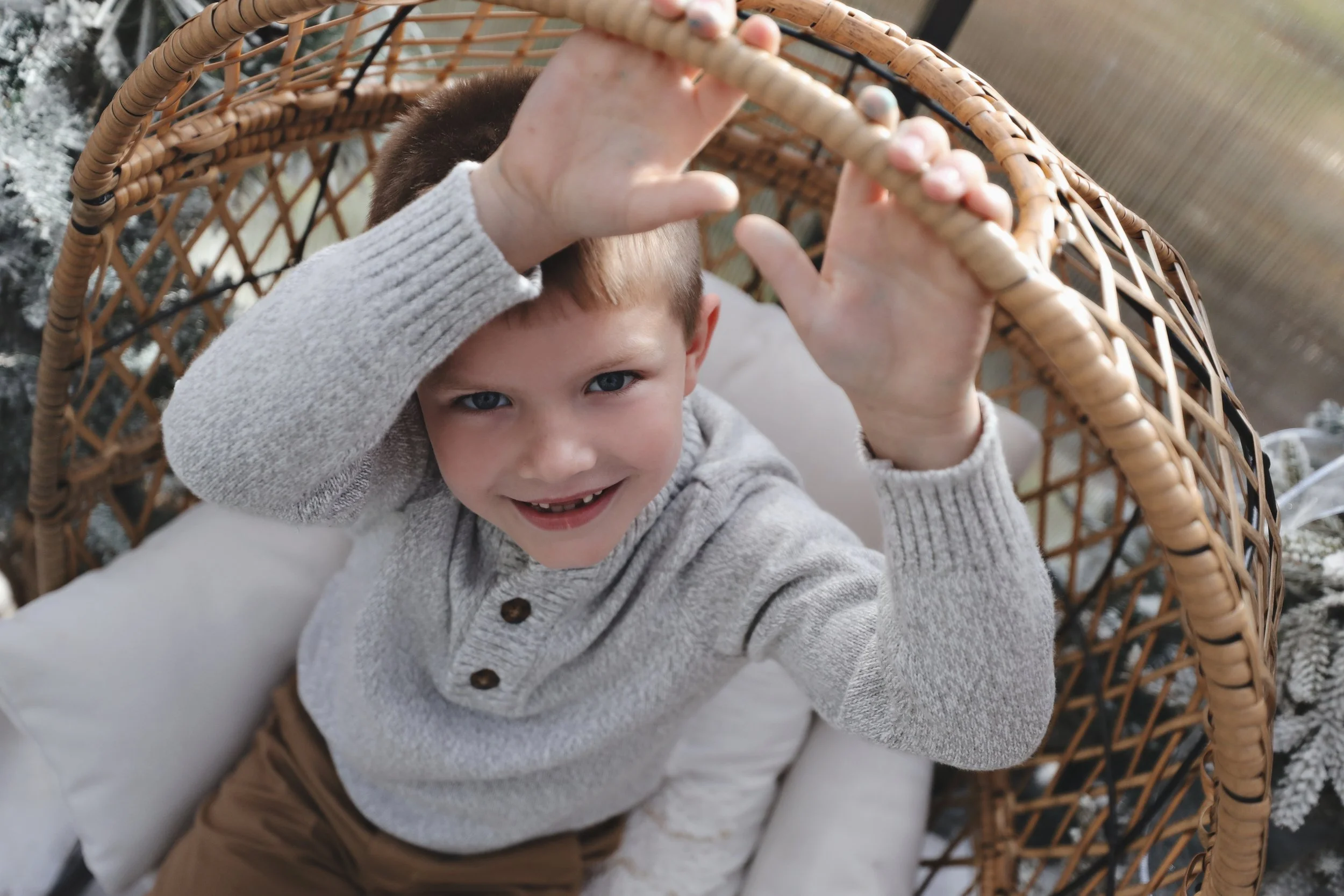 A young boy with short blond hair and blue eyes sitting in a wicker chair, smiling, wearing a grey sweater and brown pants, looking up at the camera with his hands touching the top of the chair.