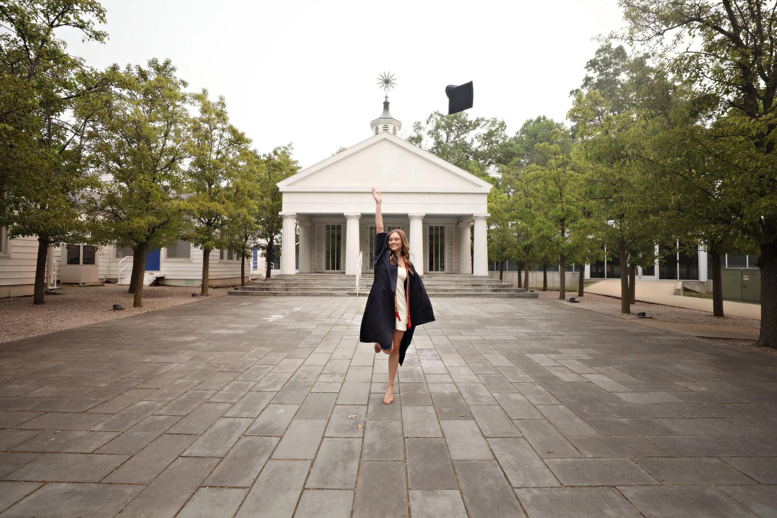 A woman in a graduation gown and cap celebrating outdoors on a paved area in front of a white building with columns, throwing her cap into the air.