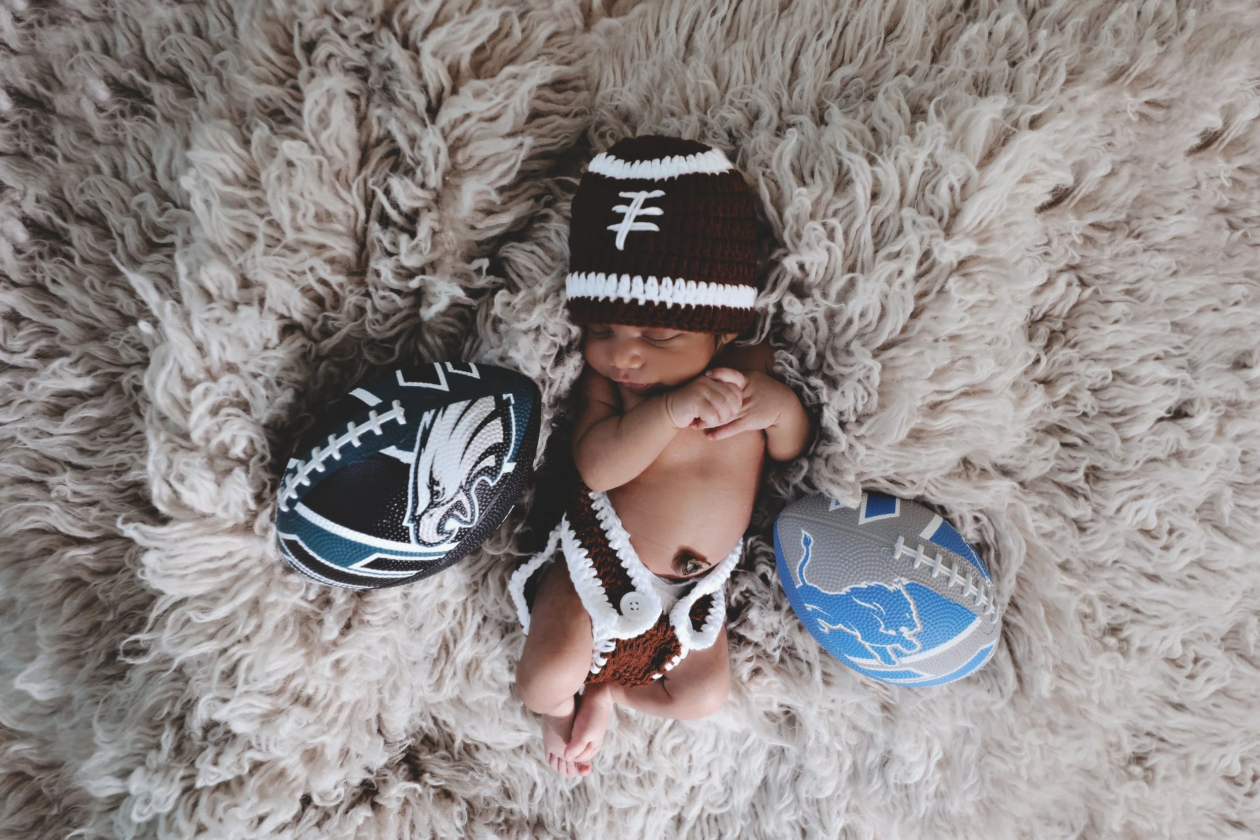 A newborn baby wearing a brown and white crochet hat, lying on a fluffy beige blanket, with two footballs on either side, one with a Philadelphia Eagles design and the other with a blue and gray design.