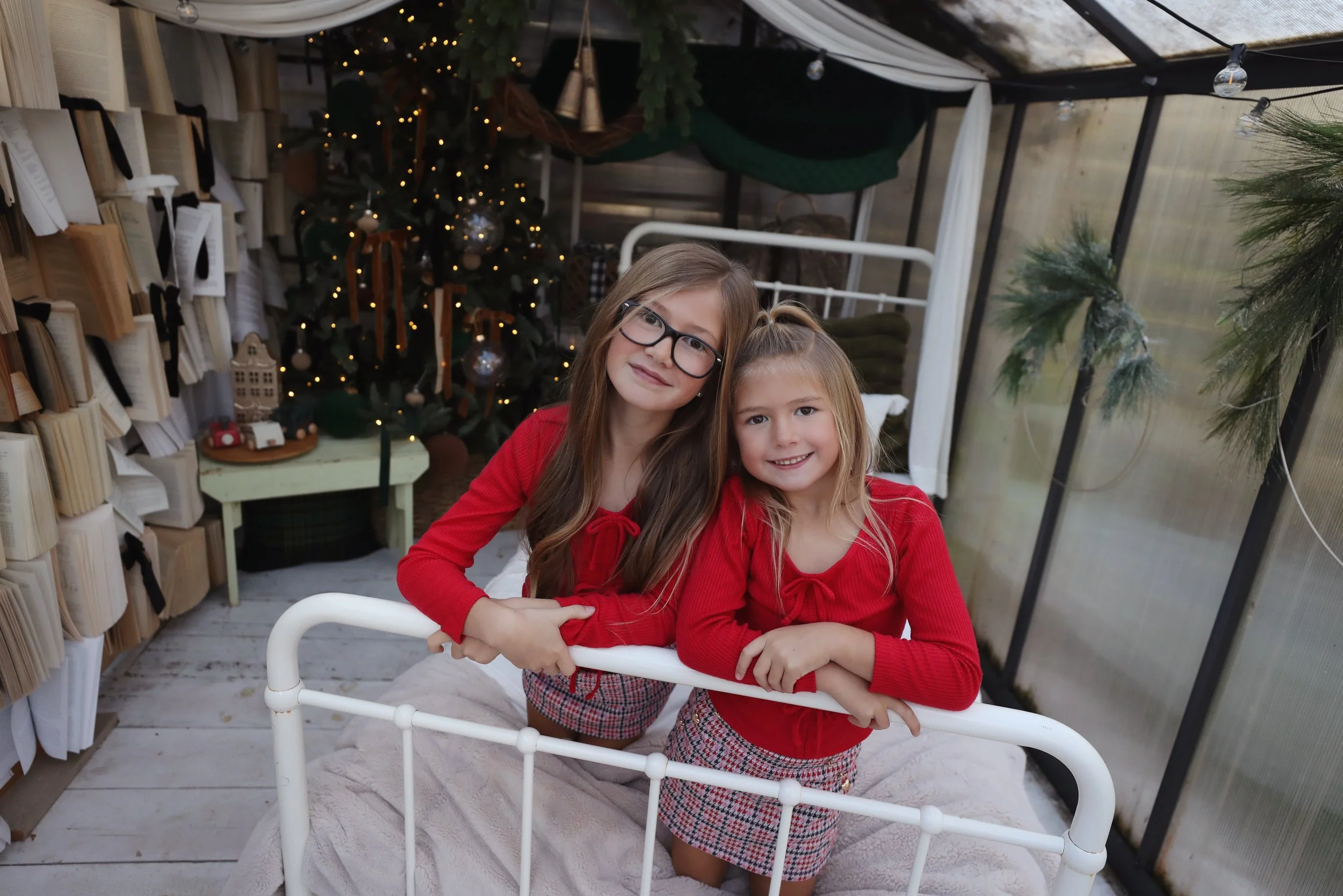 Two young girls with long hair and glasses, wearing red tops and patterned skirts, are leaning on a white bed frame in a decorated indoor space with a Christmas tree, ornaments, and books in the background.