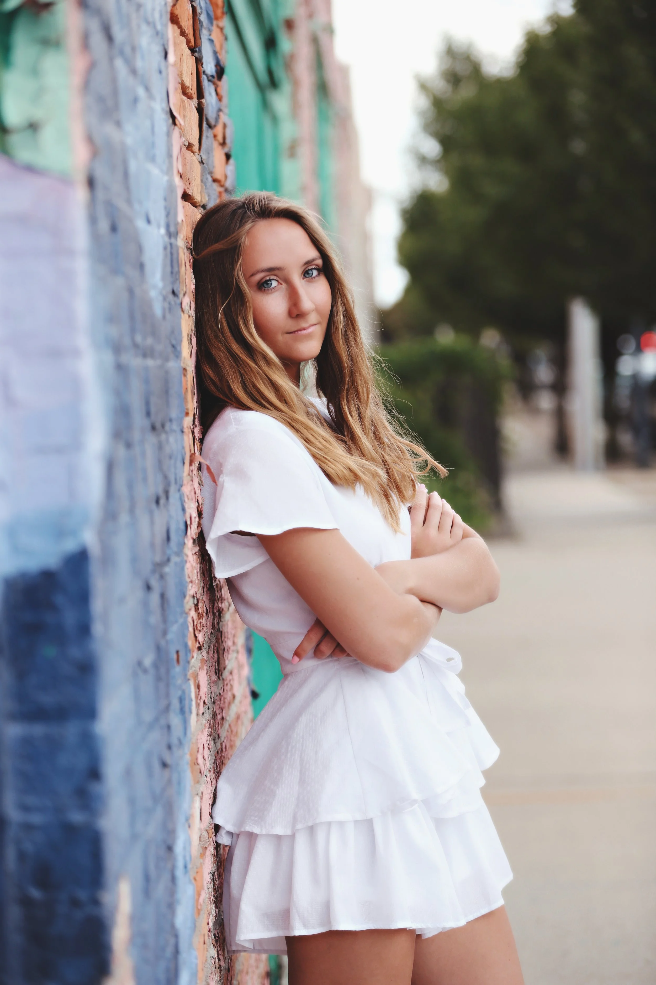 A young woman with long wavy hair leaning against a colorful brick wall with crossed arms on a sidewalk, with trees and street background.