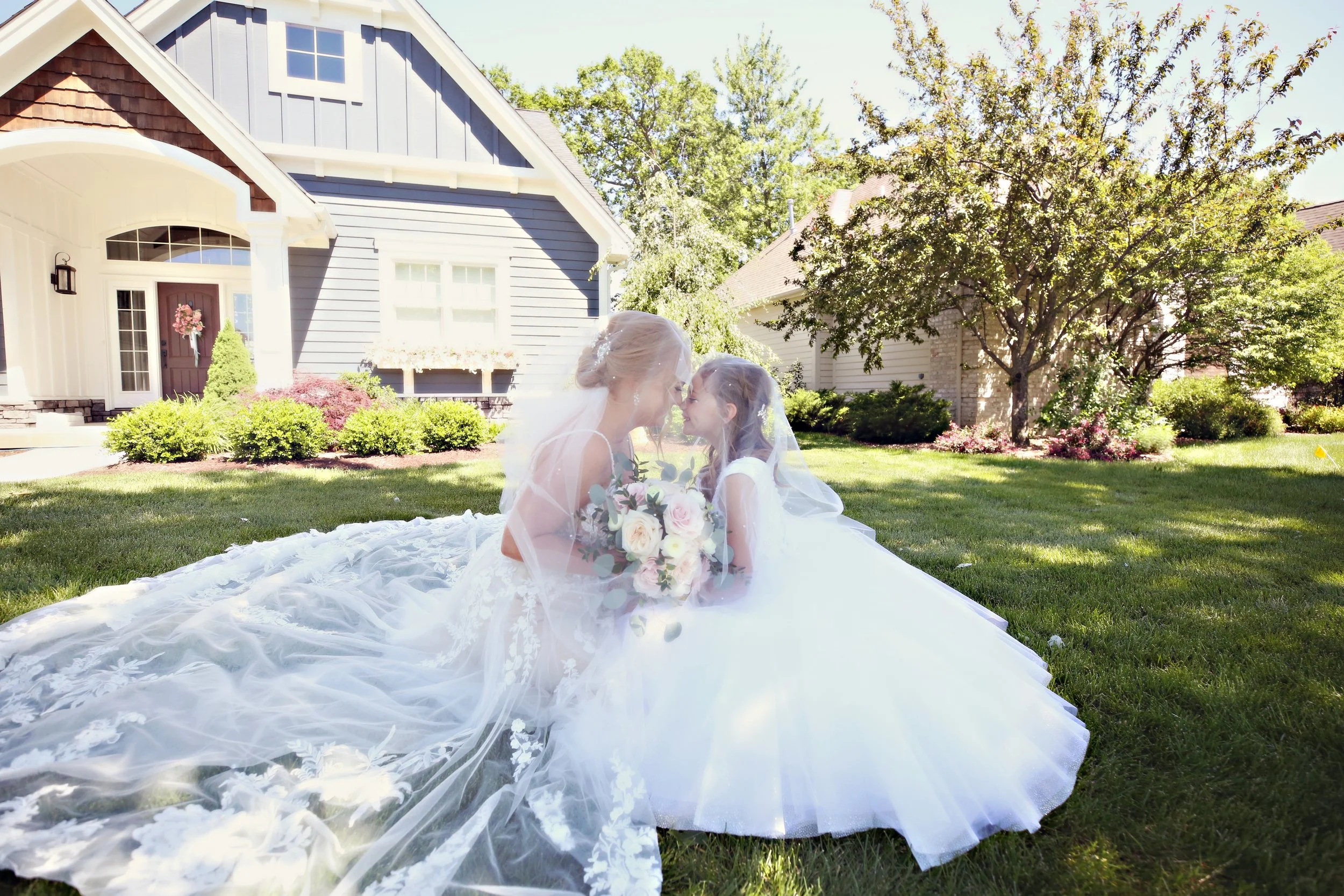 A bride in a white wedding gown and veil kneels on the grass in a suburban yard, holding a bouquet of flowers, touching foreheads with a young girl dressed in a white dress with a veil, under a tree on a sunny day.