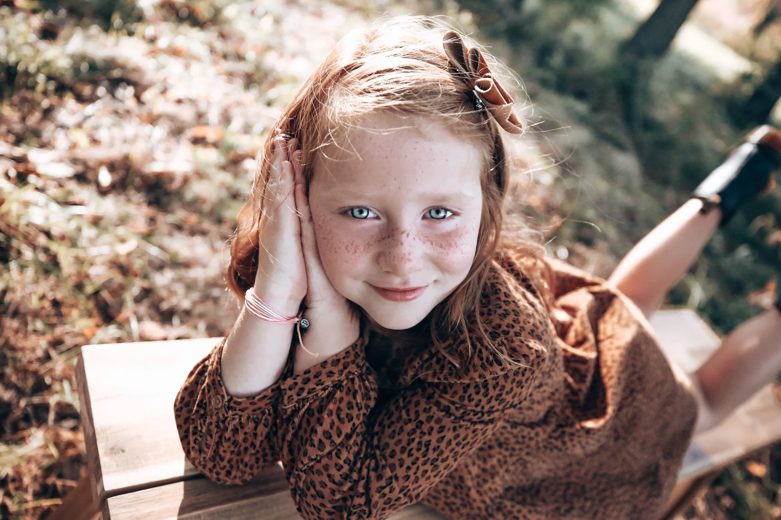 A young girl with red hair, freckles, and blue eyes lying on her stomach on a wooden surface outdoors, looking up at the camera with a smile.